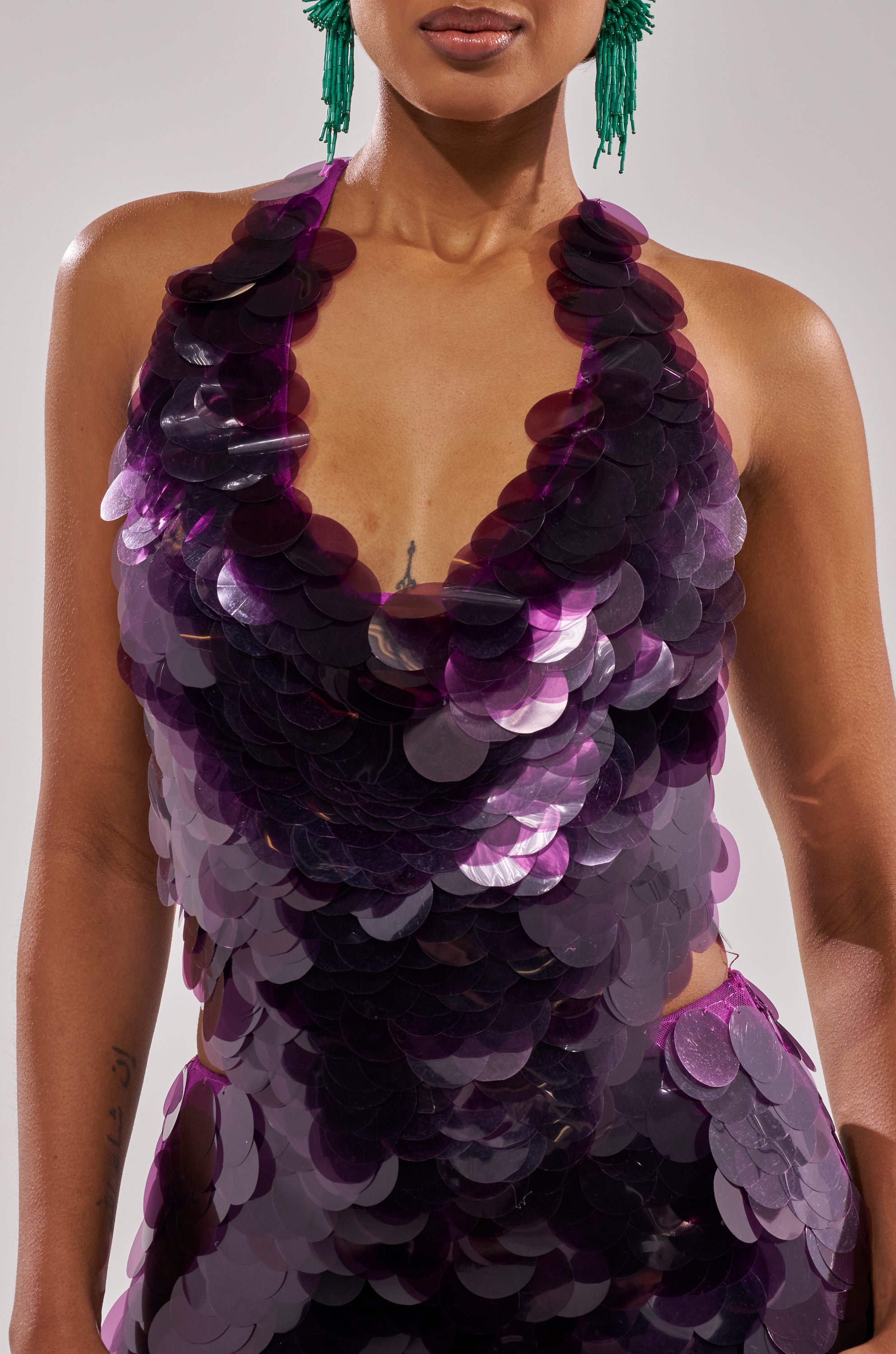 A person wears the LOVE BOMB SEQUIN BLOUSE, a shiny purple halter-style top with a deep V-neckline, paired with green statement earrings and showcasing an upper arm tattoo.