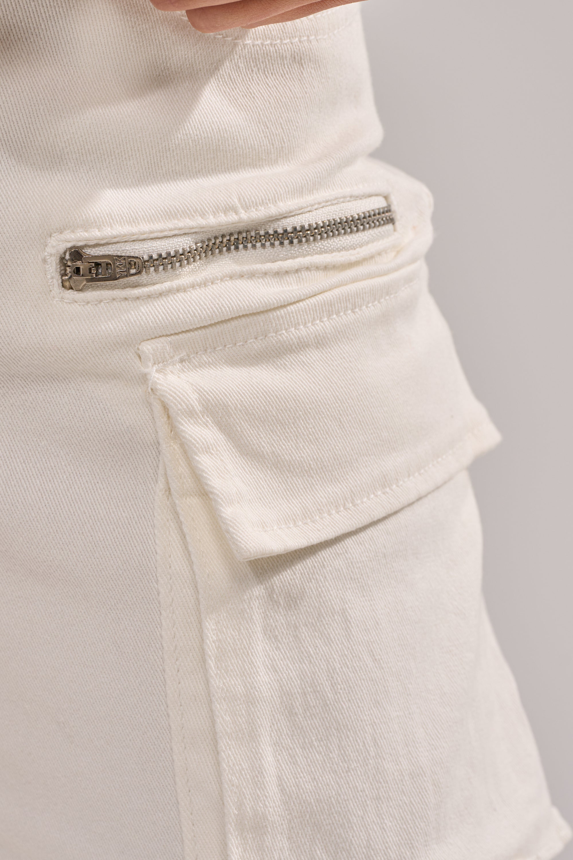 Close-up of IT GETS EASIER STACKED CARGO PANT IN WHITE, showcasing a silver zipper above a rectangular flap pocket with visible stitching. 9