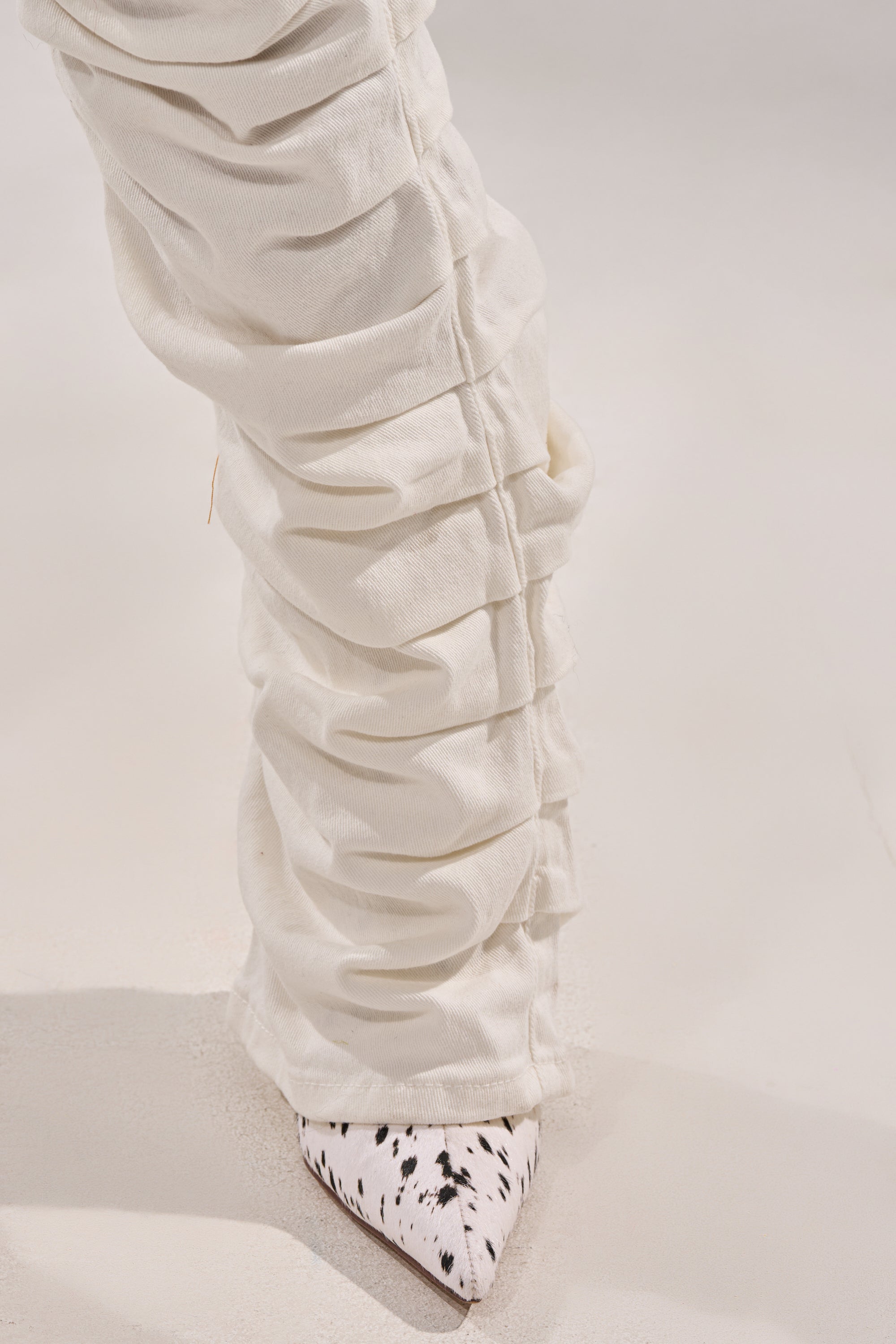 A close-up of a person wearing the IT GETS EASIER STACKED CARGO PANT IN WHITE over pointed-toe shoes with a white base and black speckles, standing on a light surface. 10