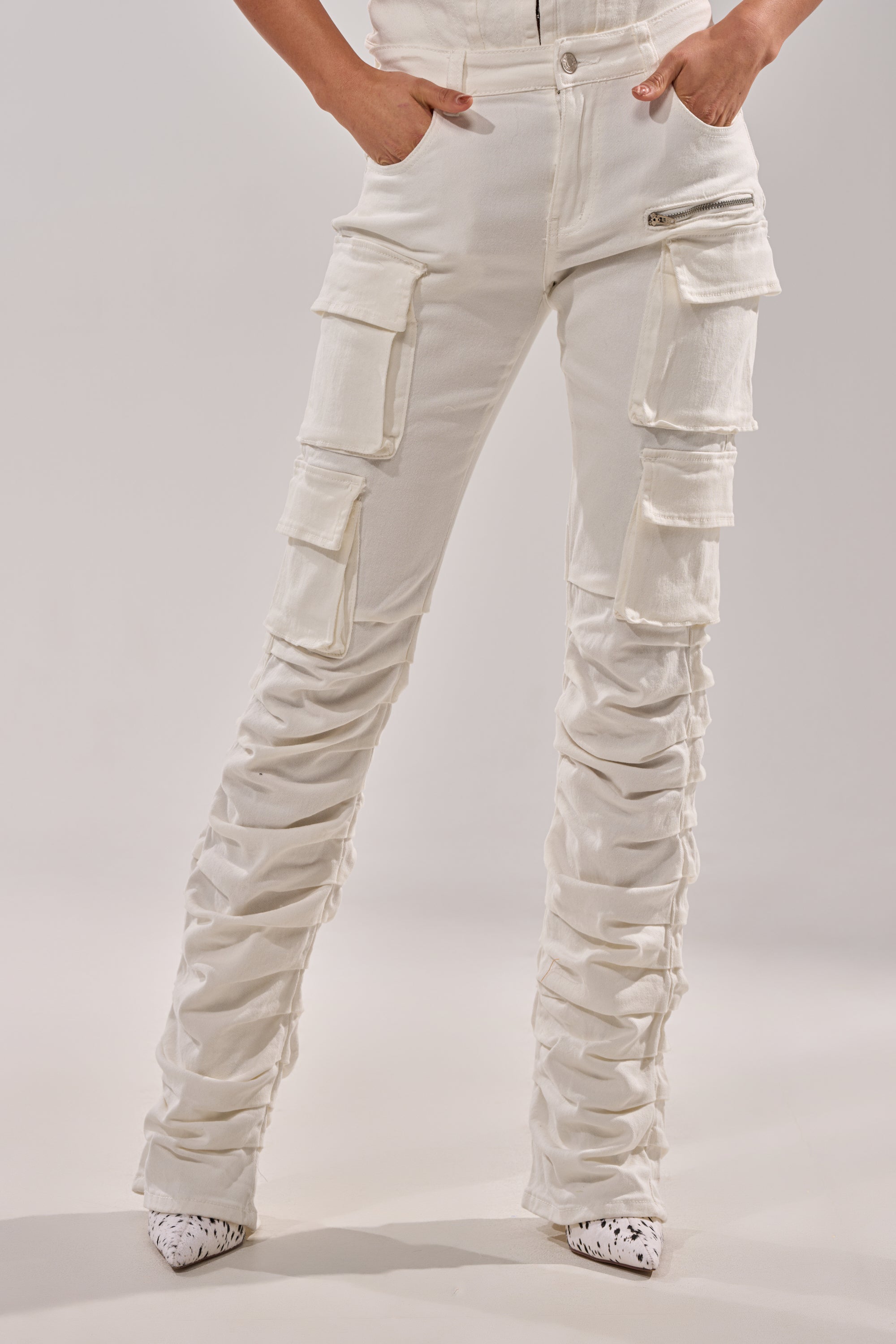 A person wears IT GETS EASIER STACKED CARGO PANT IN WHITE, featuring multiple pockets and ruched legs, paired with pointed white shoes with a black splatter pattern. Hands are in their pockets; torso is not visible. 1