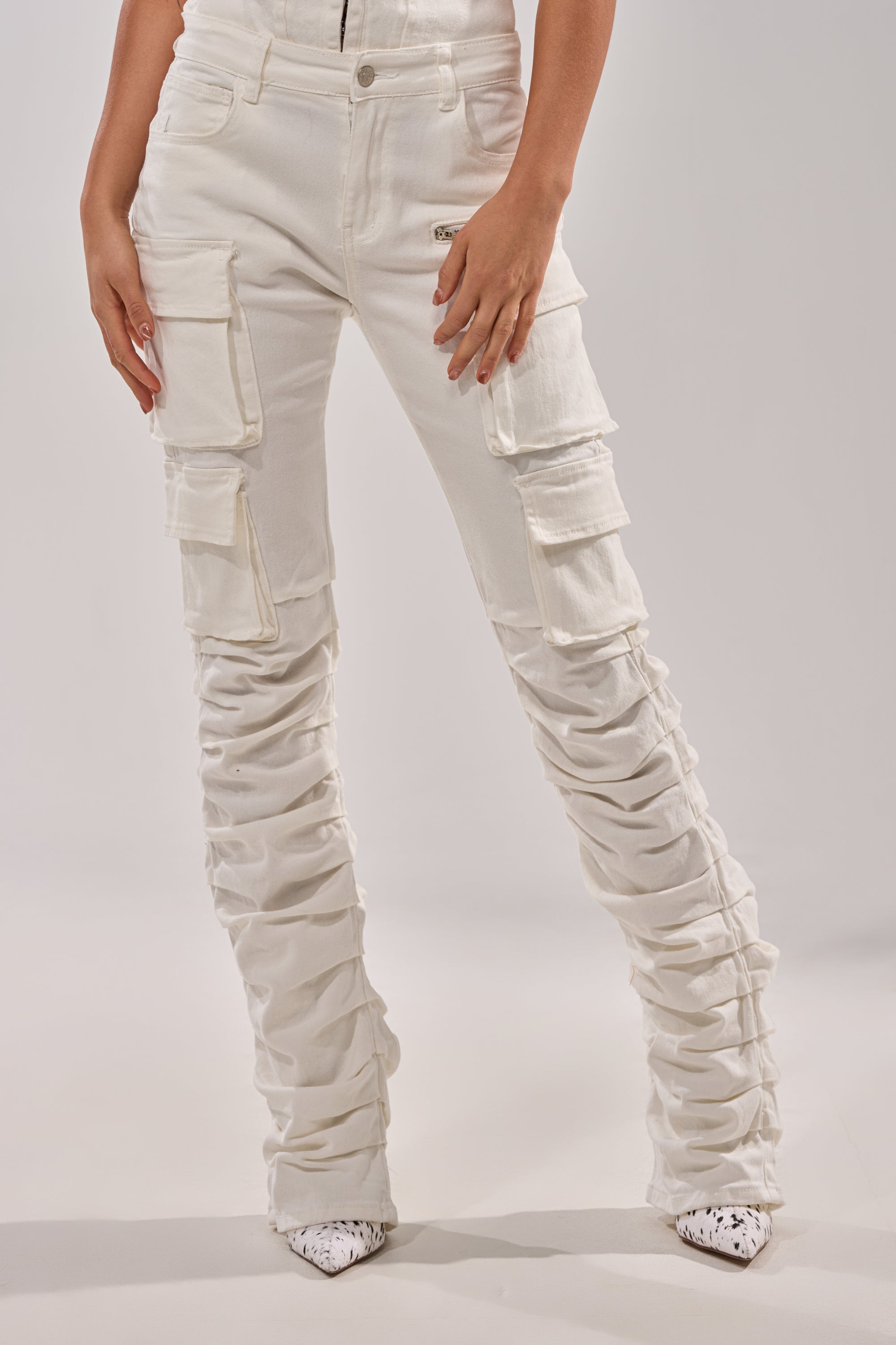 Someone stands against a plain light background wearing the IT GETS EASIER STACKED CARGO PANT IN WHITE, featuring a skinny-leg silhouette and ruched leg detailing, paired with pointed white shoes. 2