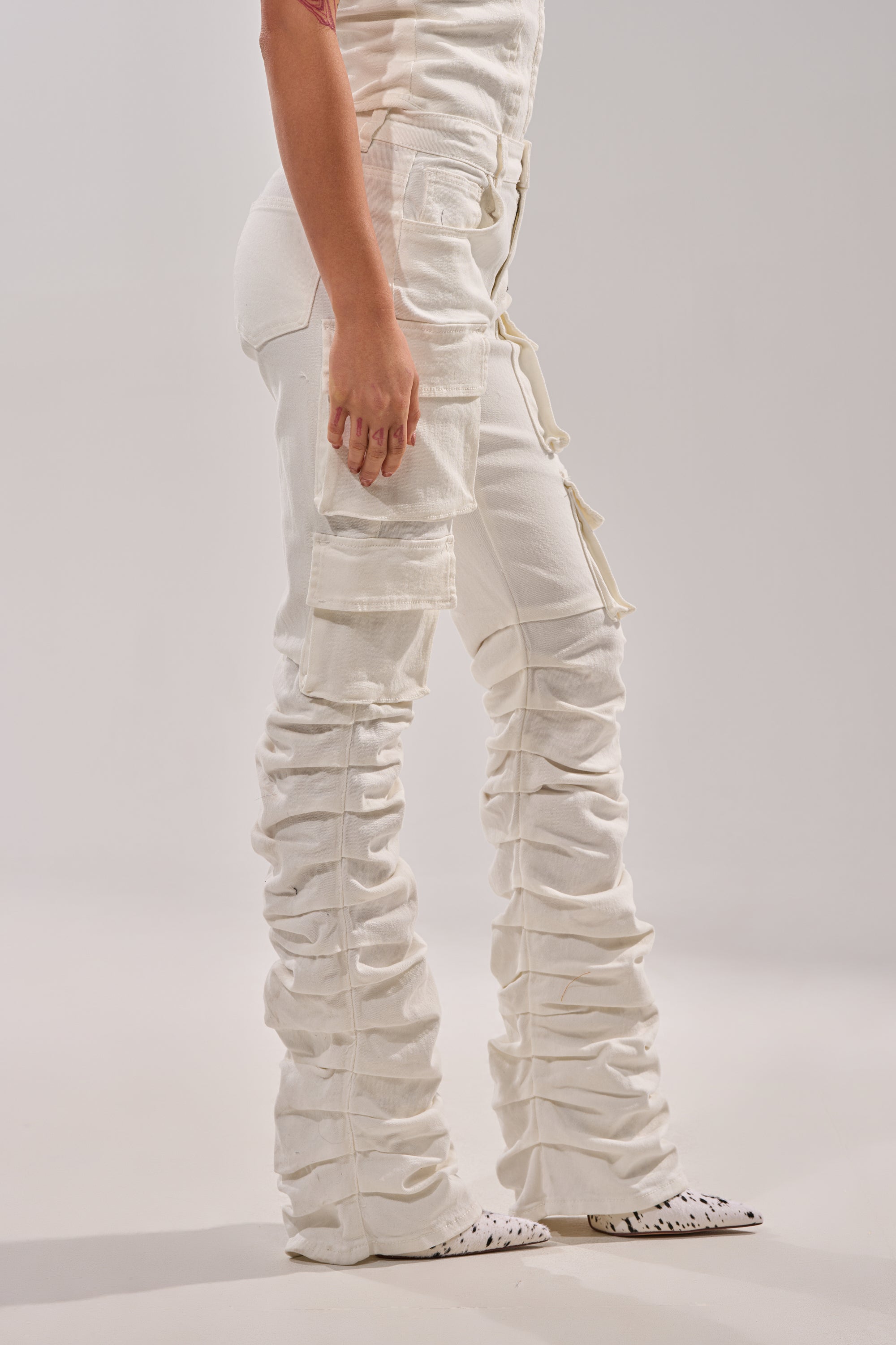 A person stands in profile wearing the IT GETS EASIER STACKED CARGO PANT IN WHITE, featuring ruched skinny legs and multiple pockets, paired with white pointed-toe shoes against a plain, light background. 3