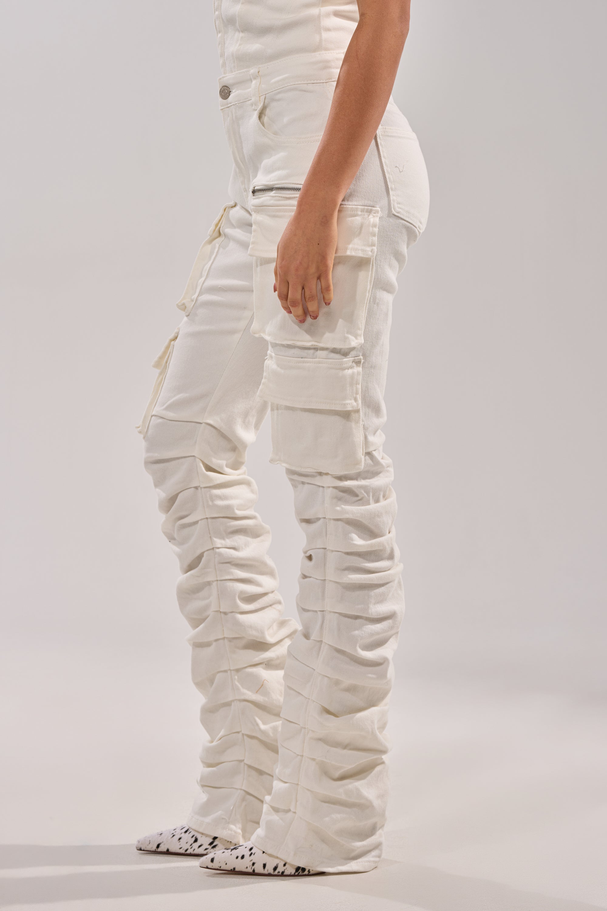 Only the lower body is visible, wearing the IT GETS EASIER STACKED CARGO PANT IN WHITE with ruched skinny legs and multiple cargo pockets, paired with white pointed-toe shoes featuring black spots against a plain background. 4