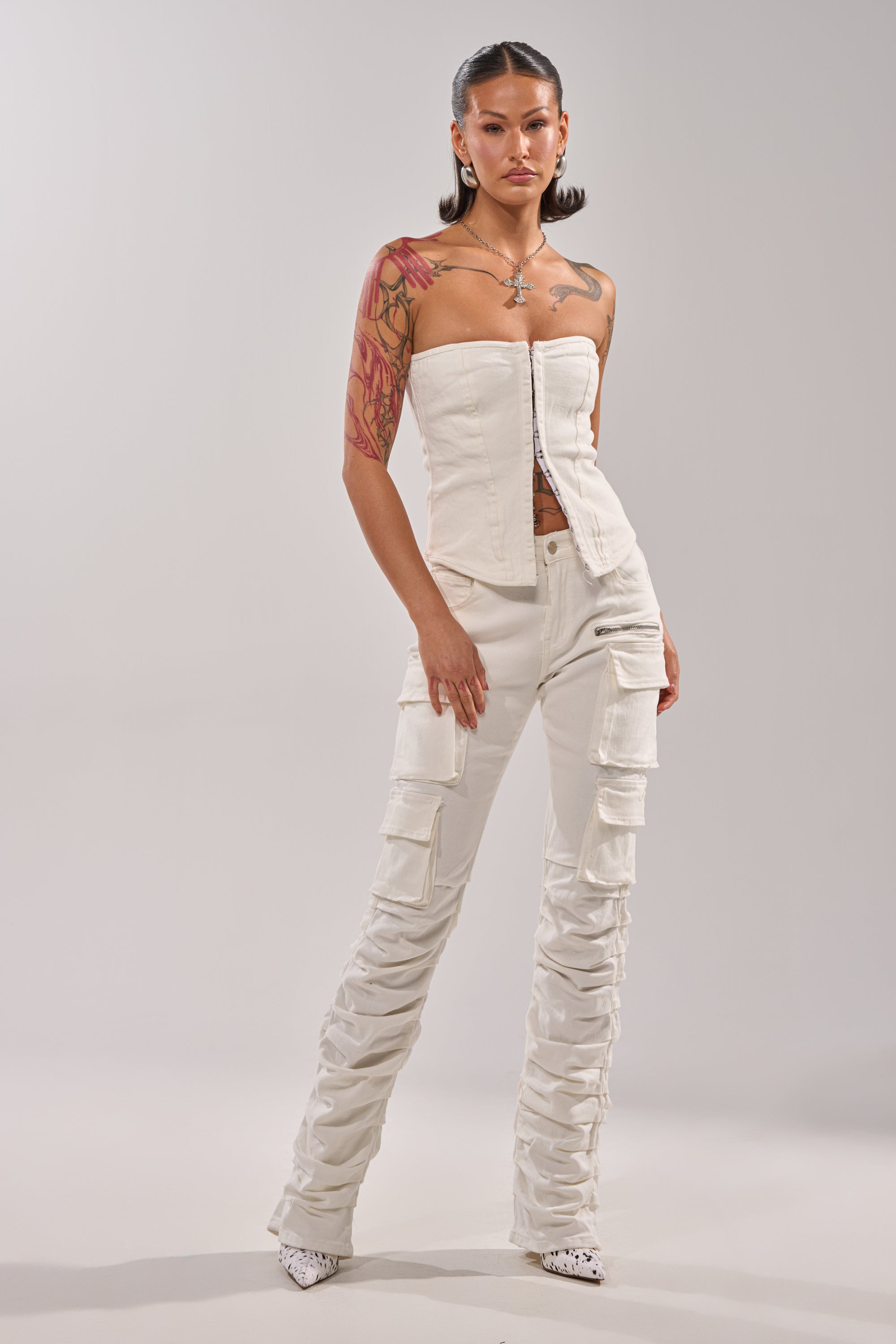 A woman stands confidently in a white strapless corset top and the IT GETS EASIER STACKED CARGO PANT IN WHITE, featuring multiple pockets. She has slicked-back hair, visible tattoos, white pointed shoes, and silver jewelry against a plain background. 6