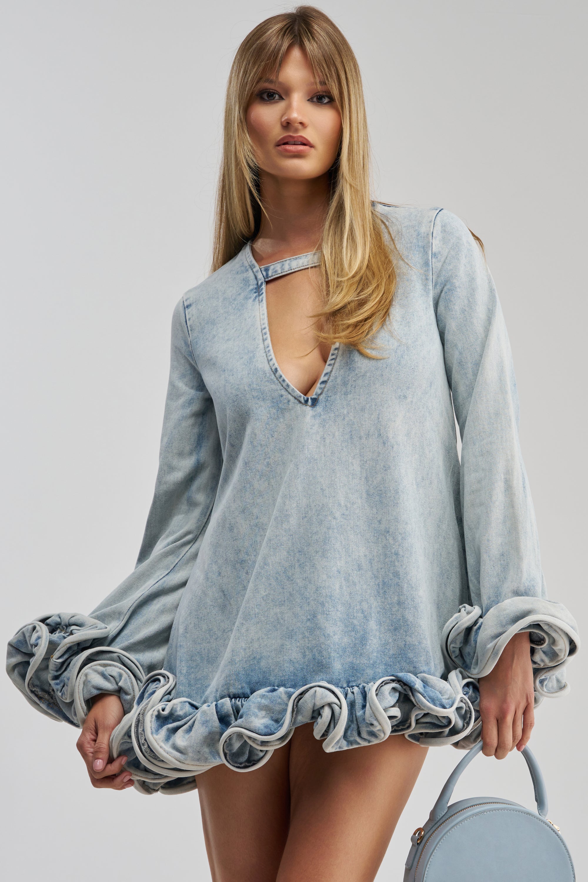 A woman wears the GIRLY GIRL MINI DRESS IN LIGHT BLUE DENIM, featuring long puffed sleeves and a ruffled hem, paired with a matching bag as she stands against a plain light background. This chic mini dress is perfect for adding a touch of effortless style to your date night look.