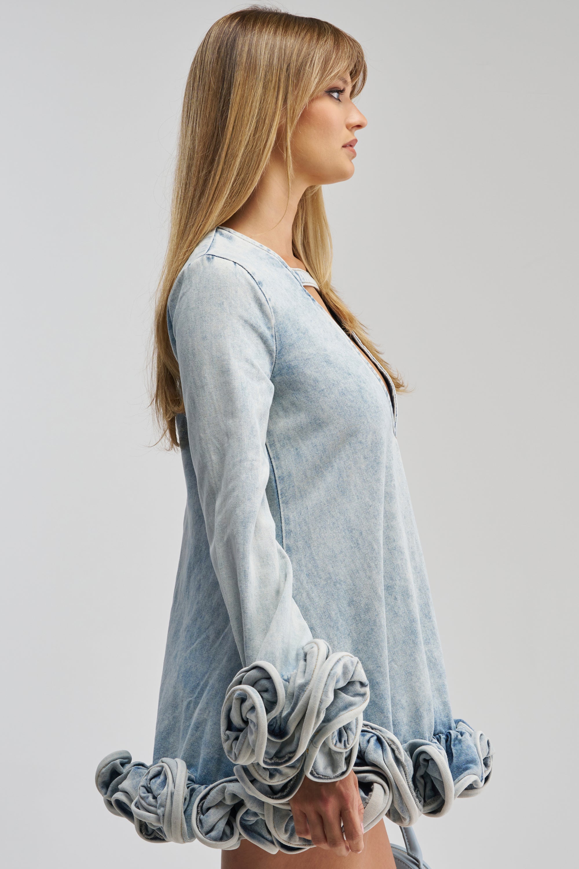 A woman in profile wears the GIRLY GIRL MINI DRESS IN LIGHT BLUE DENIM, a light wash denim mini dress featuring long sleeves and ruffled, rolled fabric trim at the cuffs and hem, standing against a plain light background. This flirty date night look brings modern edge to classic denim with its playful details.