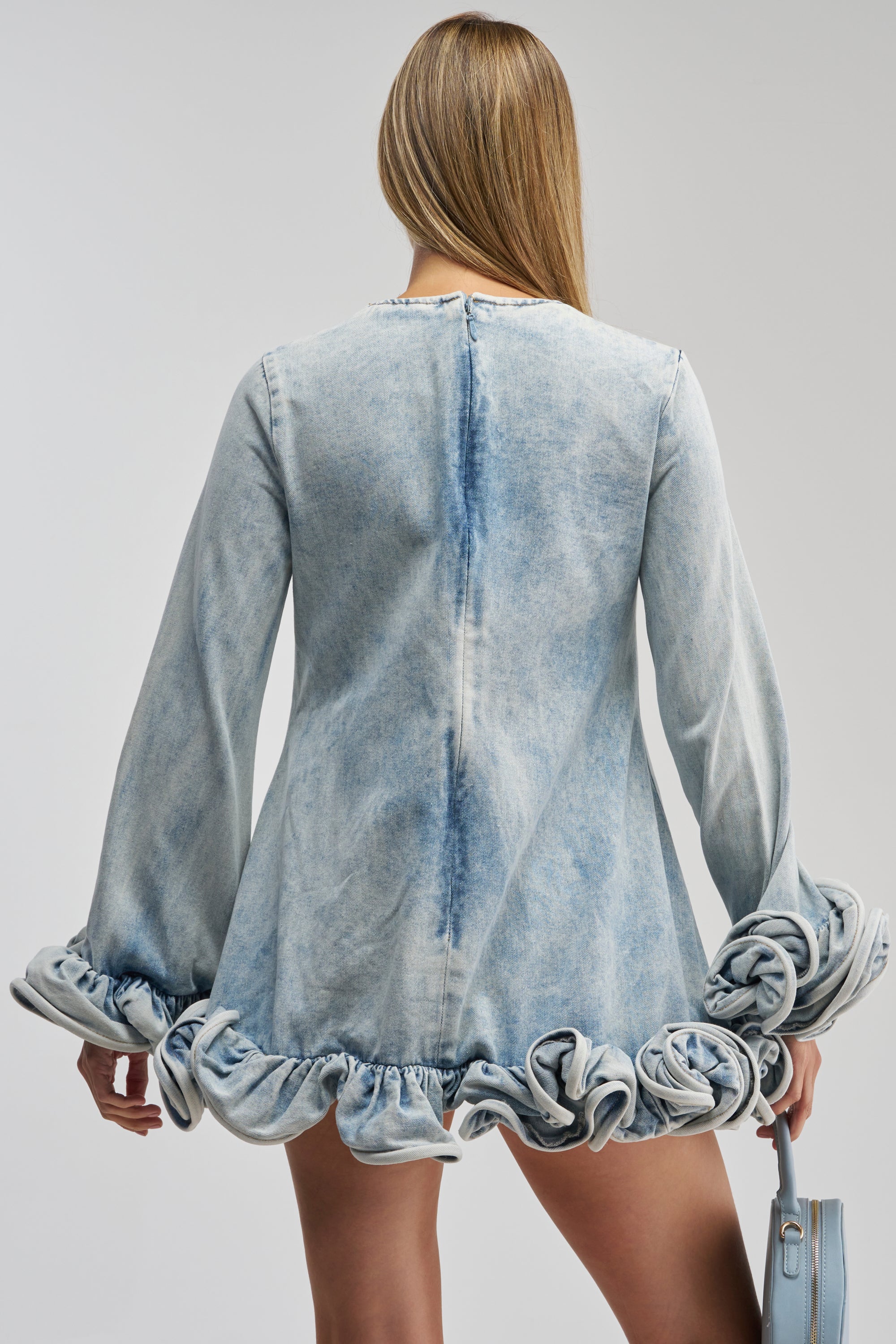 A person with long, straight hair faces away, wearing the GIRLY GIRL MINI DRESS IN LIGHT BLUE DENIM, a faded blue light wash mini dress featuring ruffled trim at the cuffs and hem, and holding a matching light blue handbag.