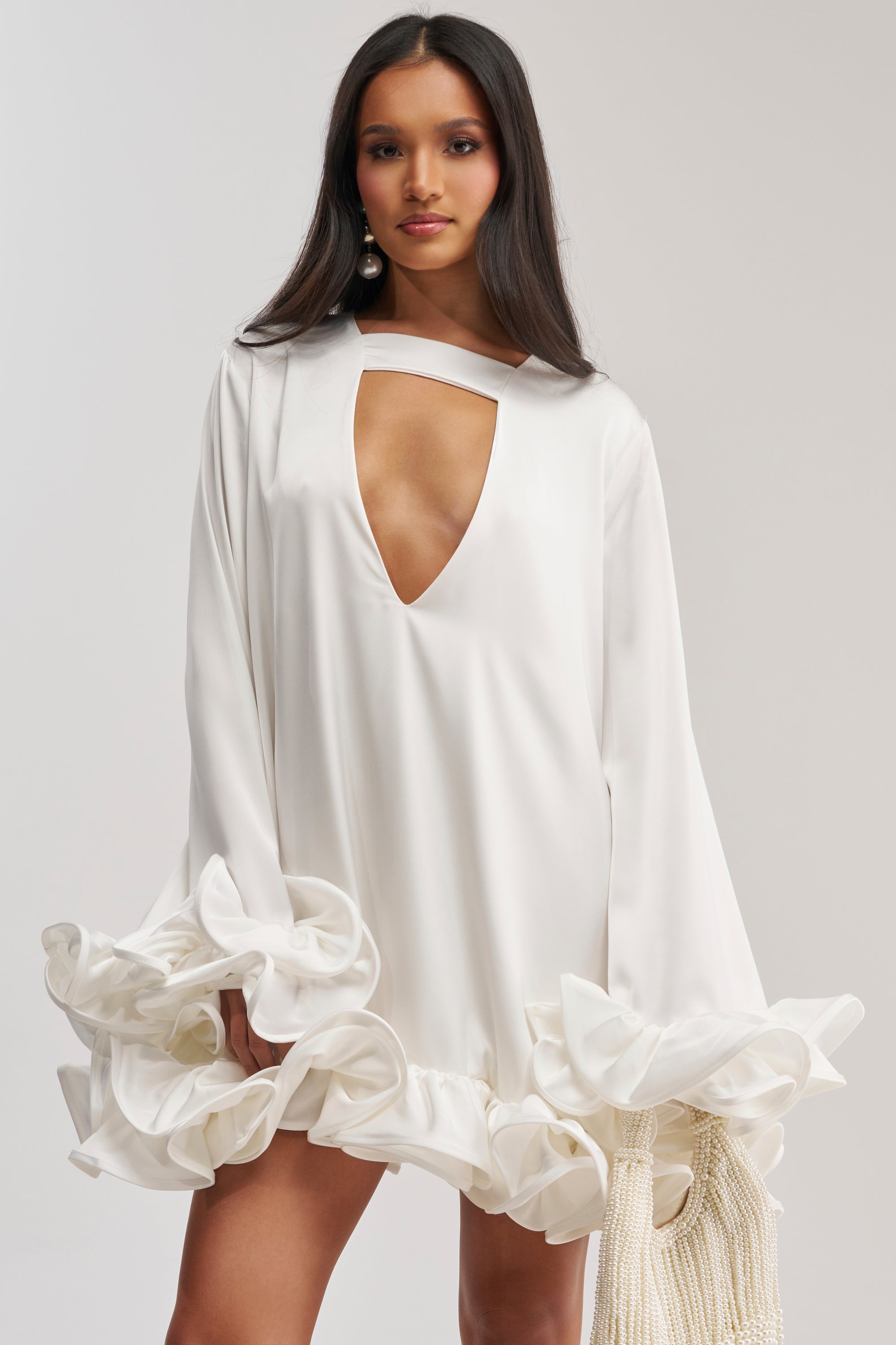 A woman wears the GIRLY GIRL SATIN MINI DRESS IN WHITE, a loose mini dress with a deep V-neck, choker collar, and large ruffled hem. She completes the look with long dark hair and a pearl-adorned beaded handbag.