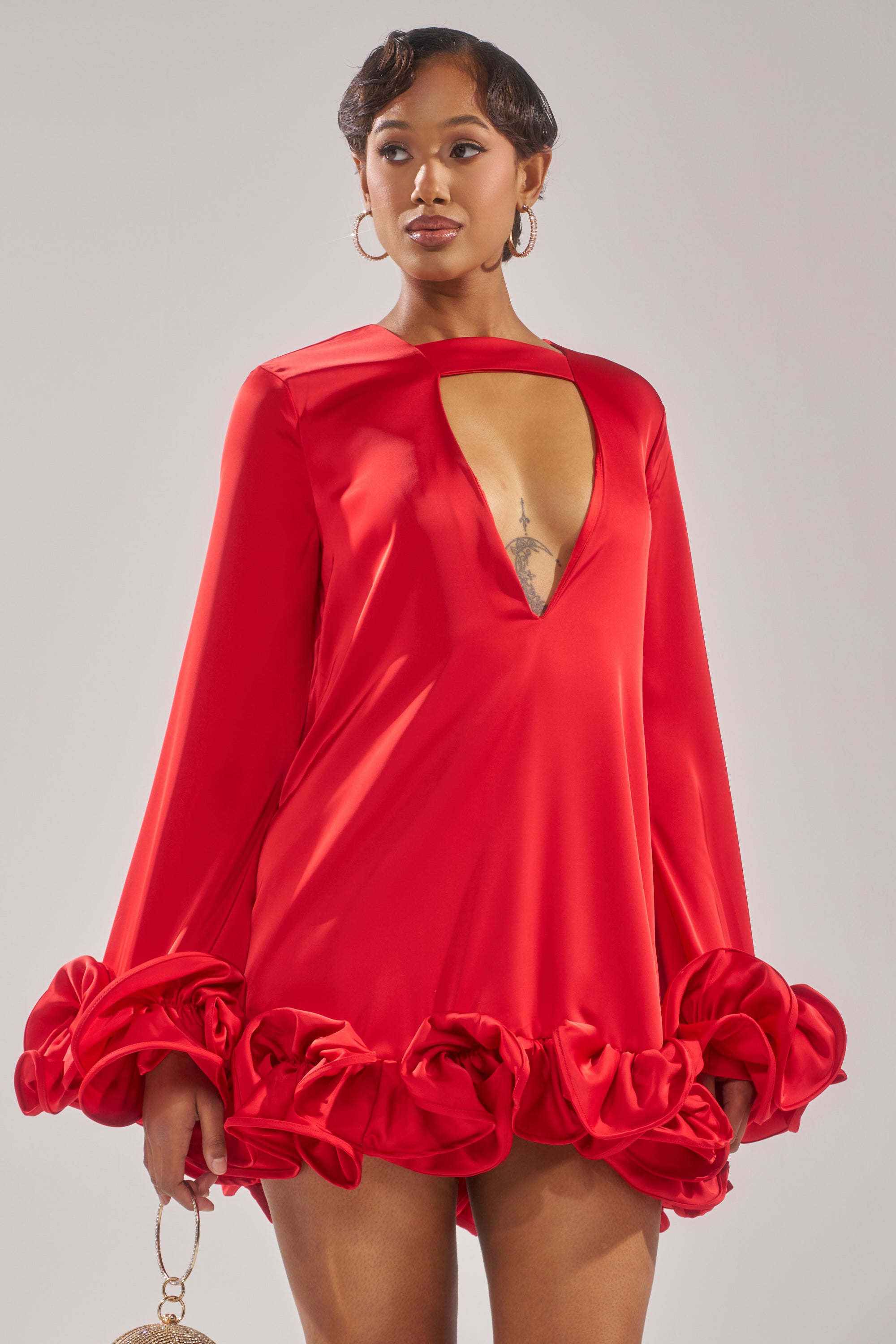 A person poses confidently against a neutral background in the GIRLY GIRL SATIN MINI DRESS, featuring vibrant red satin fabric, long ruffle sleeves, a deep V-neckline, and ruffled hem and cuffs.