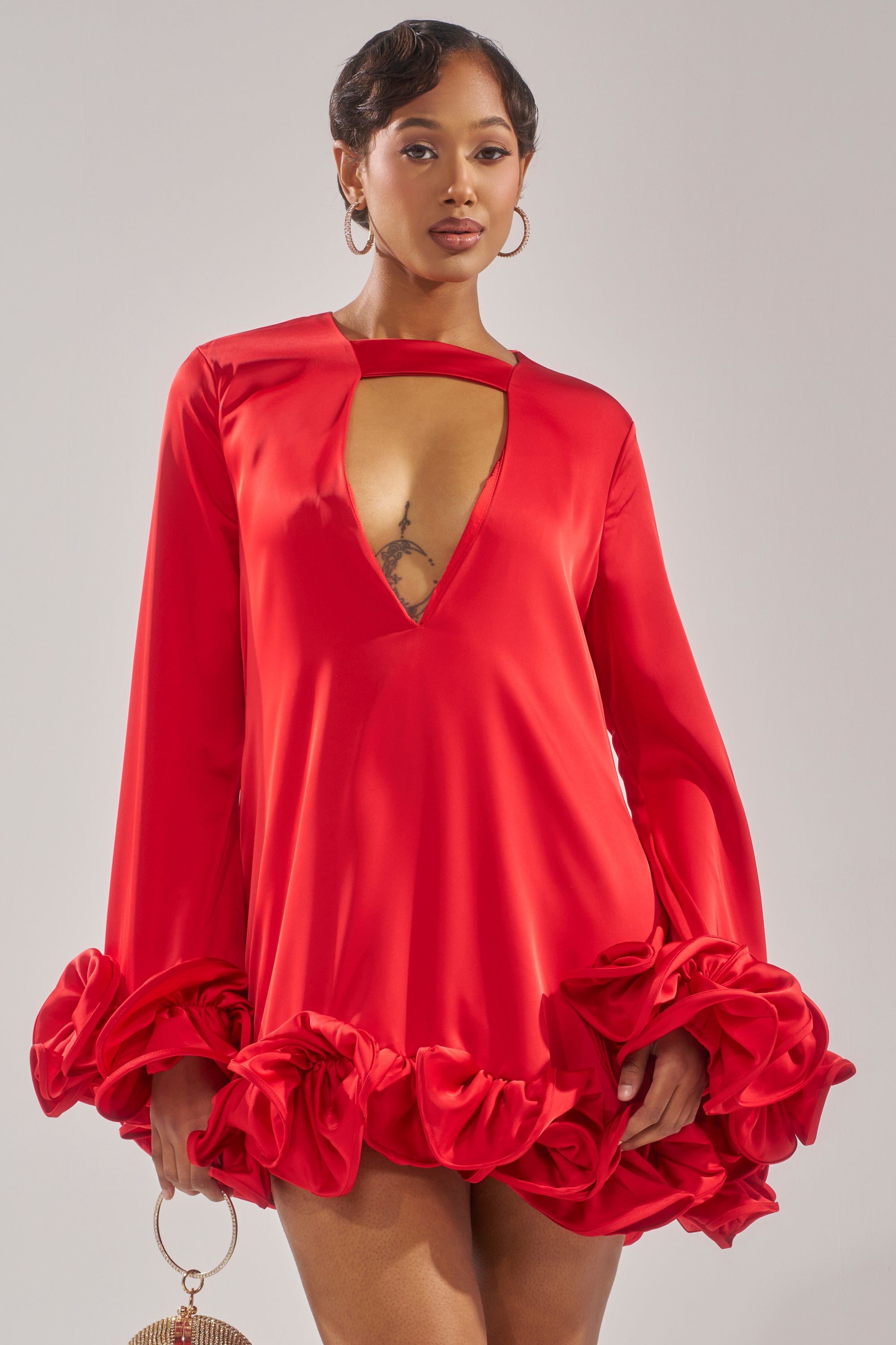 A woman stands confidently in the GIRLY GIRL SATIN MINI DRESS, featuring a bright red hue, ruffle detailing, and a deep V neckline. She holds a small round handbag, her short hair styled and accessorized with large hoop earrings.