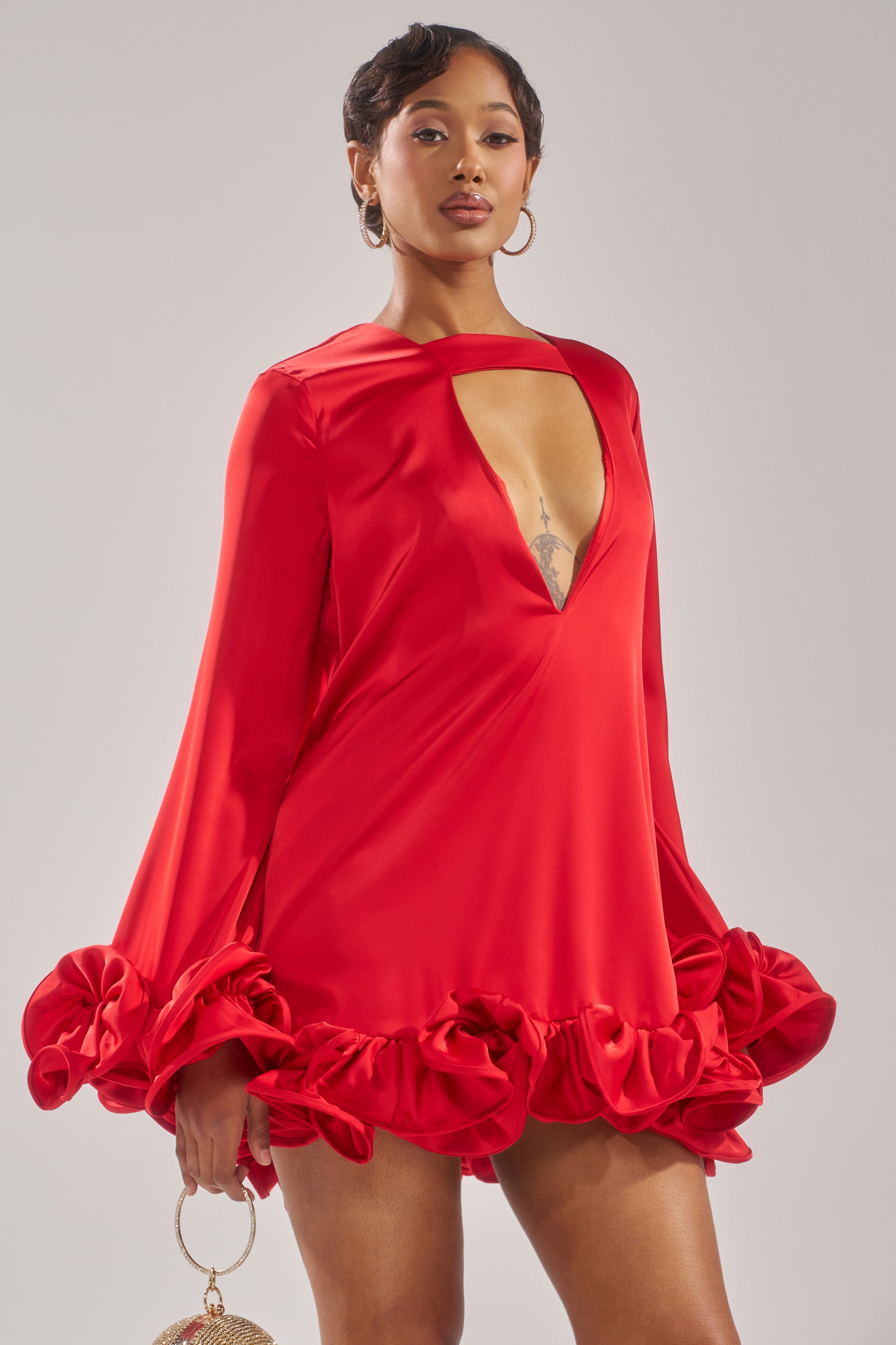 A woman poses confidently in the GIRLY GIRL SATIN MINI DRESS, a bold red mini with a deep V-neck and ruffled hem, holding a small round handbag against a plain light background.