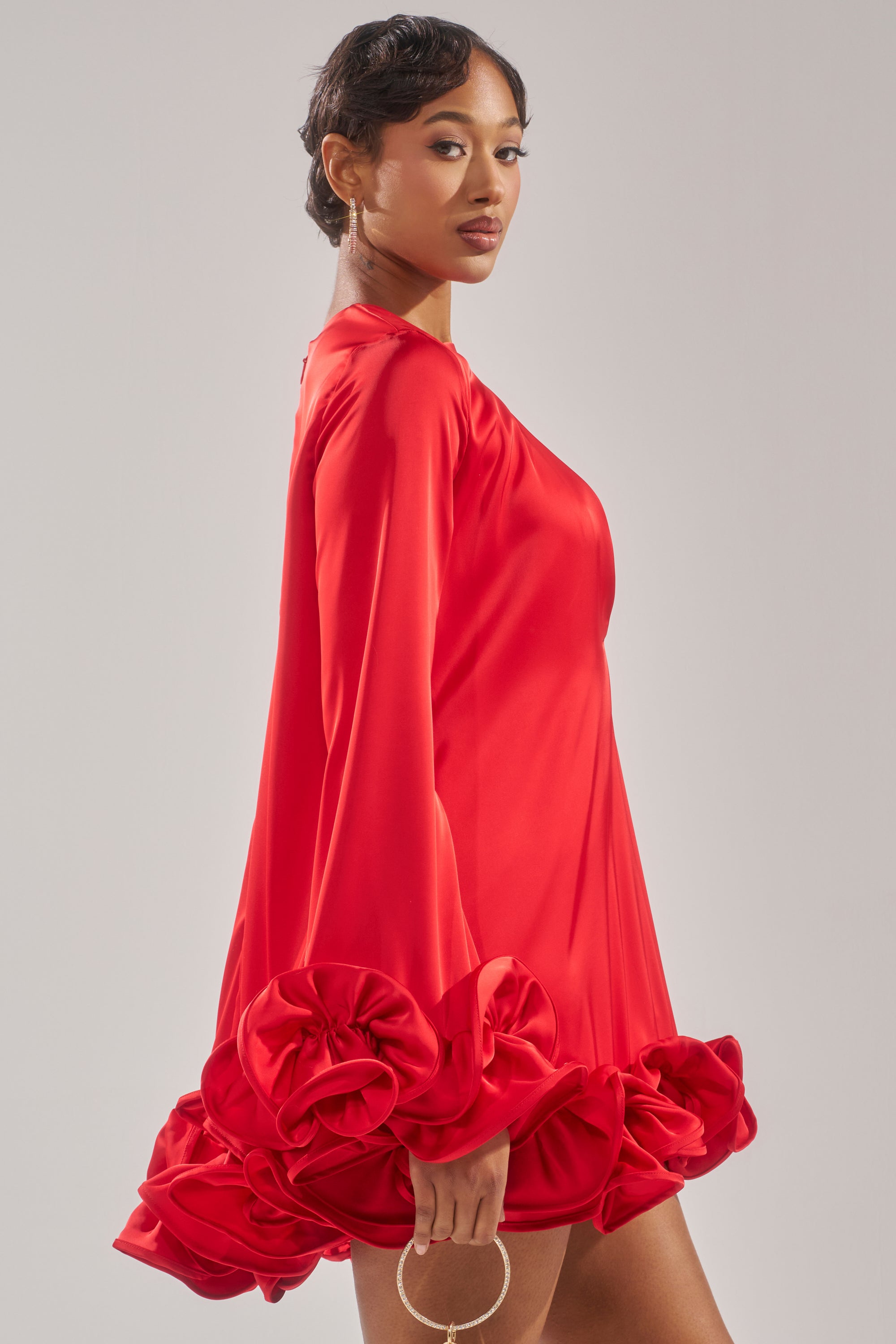 A woman wears the GIRLY GIRL SATIN MINI DRESS, a bold red mini dress with dramatic ruffle hem detailing. She holds a small circular handbag and stands sideways, looking confidently over her shoulder against a plain background.