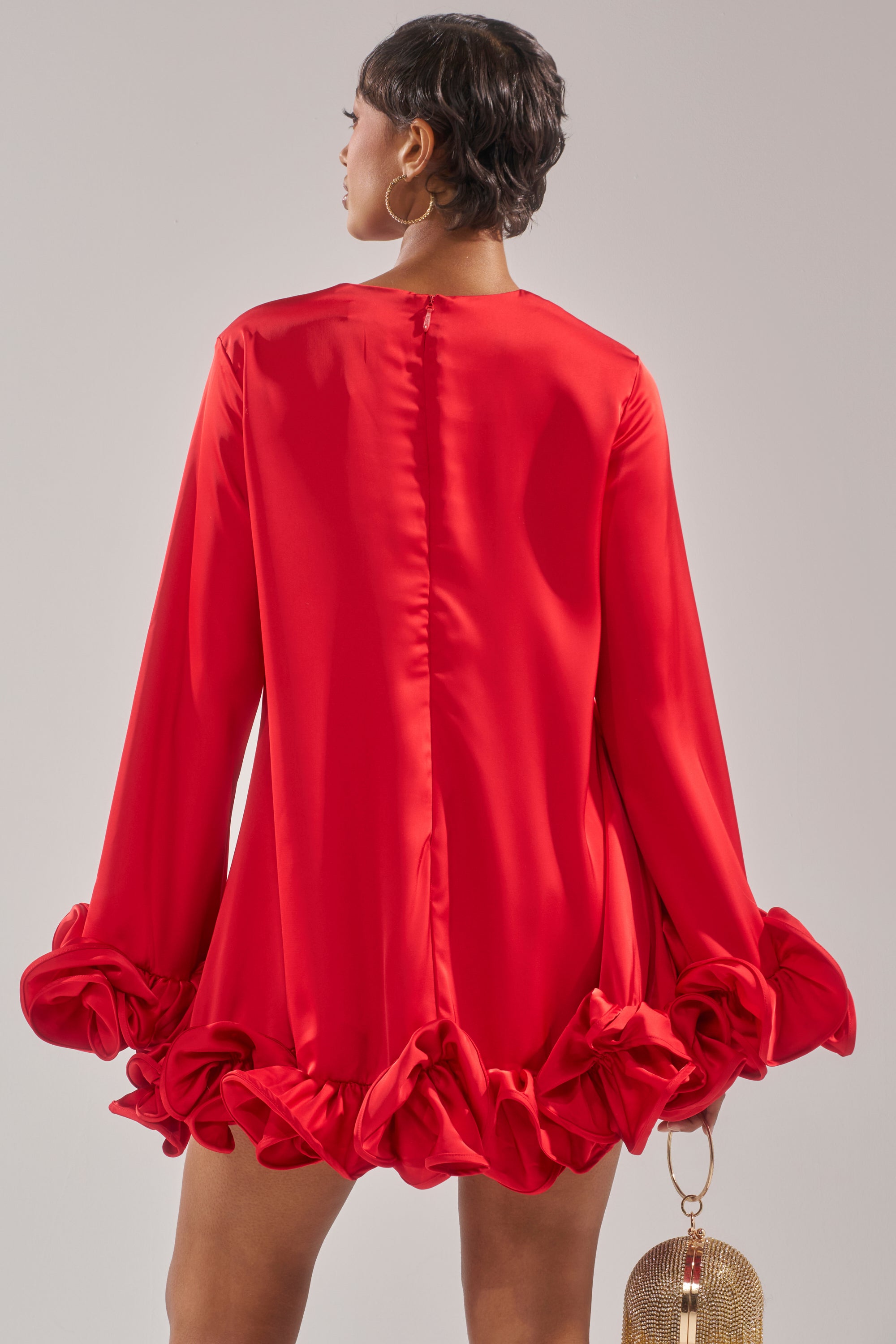 A person faces away, wearing the GIRLY GIRL SATIN MINI DRESS featuring ruffle cuffs and hem. They hold a gold circular mesh handbag, accessorized with gold hoop earrings and short dark hair.