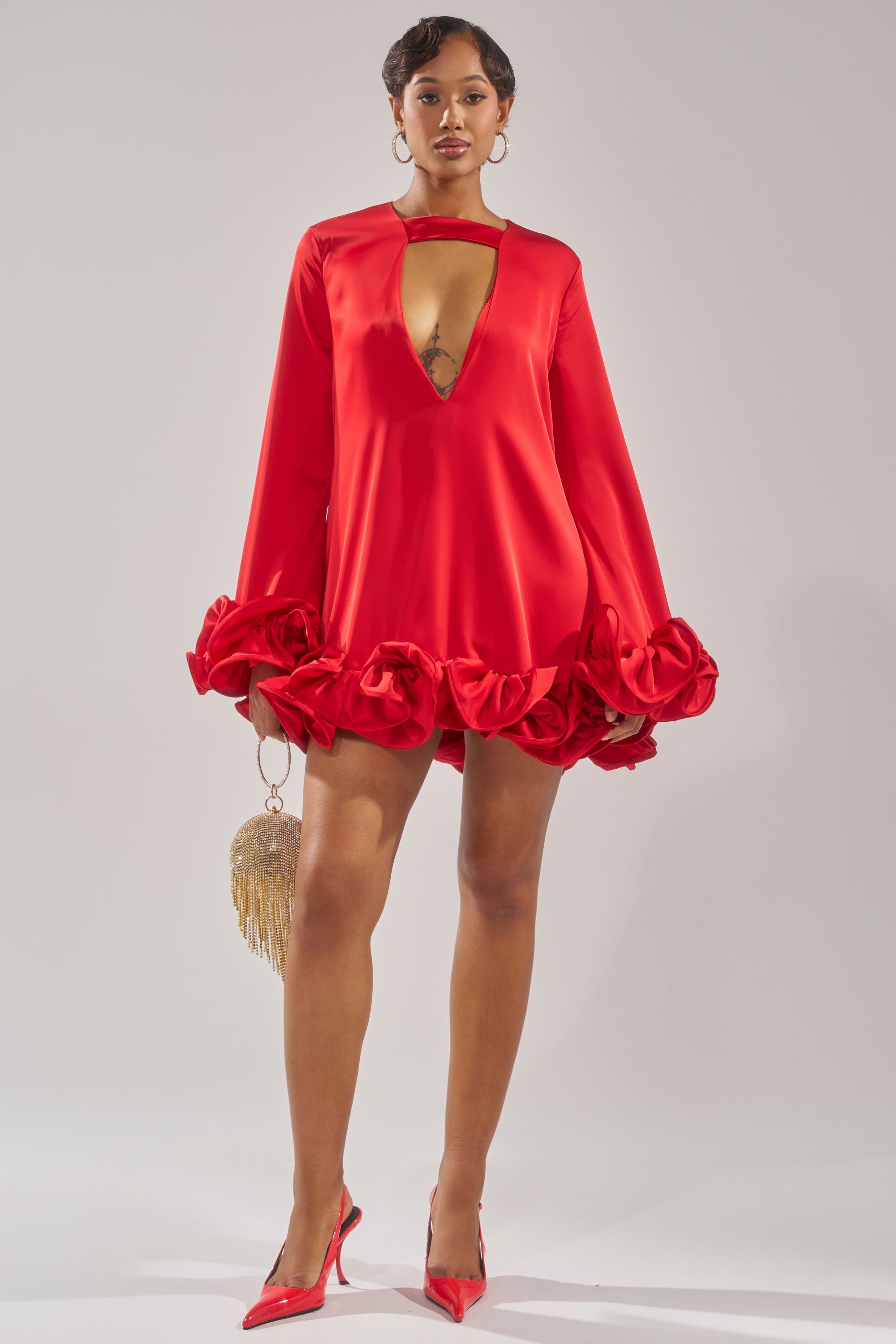 A woman stands confidently in the GIRLY GIRL SATIN MINI DRESS, a red mini with ruffle details on the hem and sleeves. She carries a gold fringe purse, wears red pointed heels, has short hair, and finishes her look with chic hoop earrings.