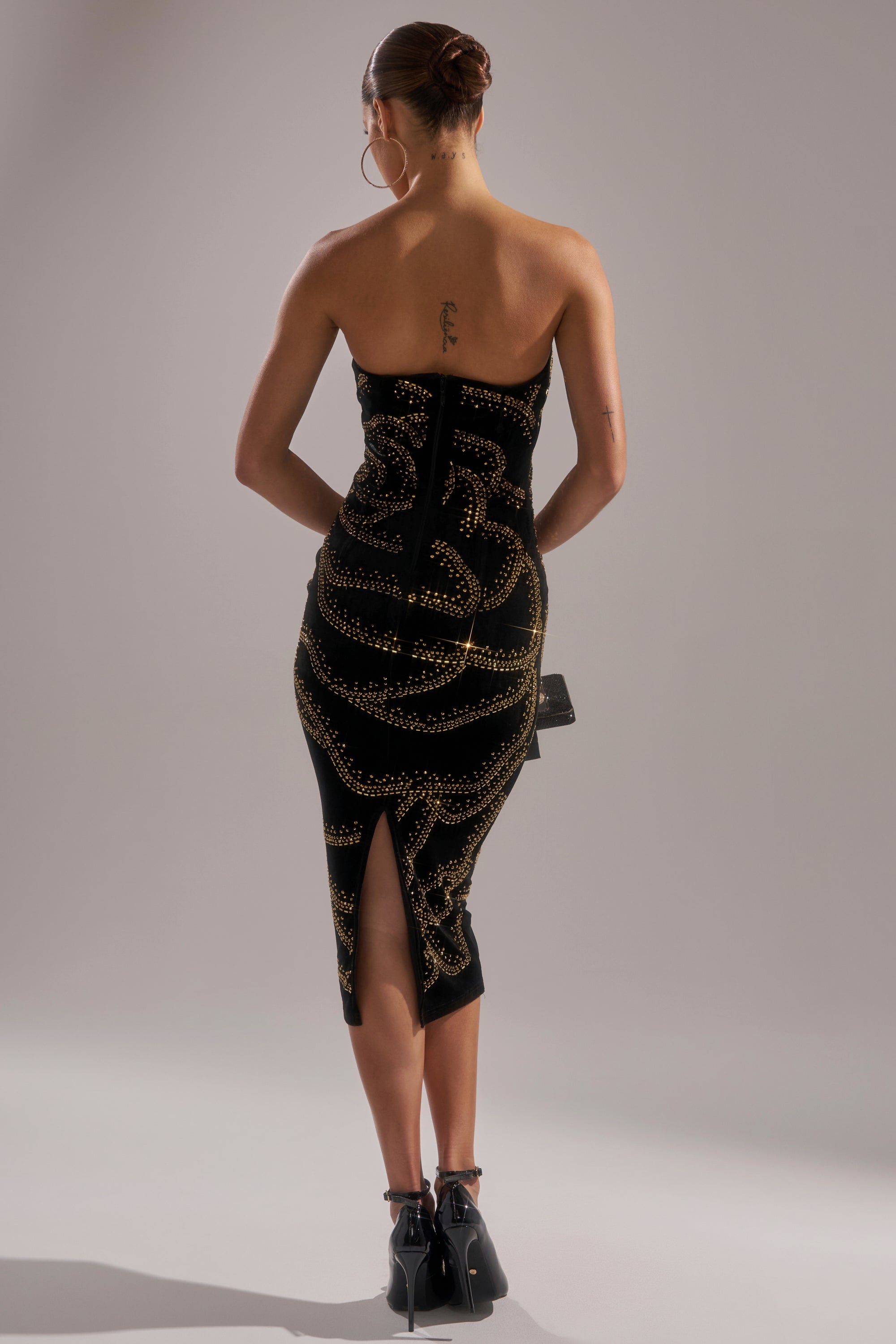 A woman stands facing away, wearing the SHIMMER IN GOLD MIDI DRESS, a strapless velvet design with gold embellishments. She pairs it with black heels and hoop earrings, her hair in a neat bun that reveals a tattoo on her upper back.