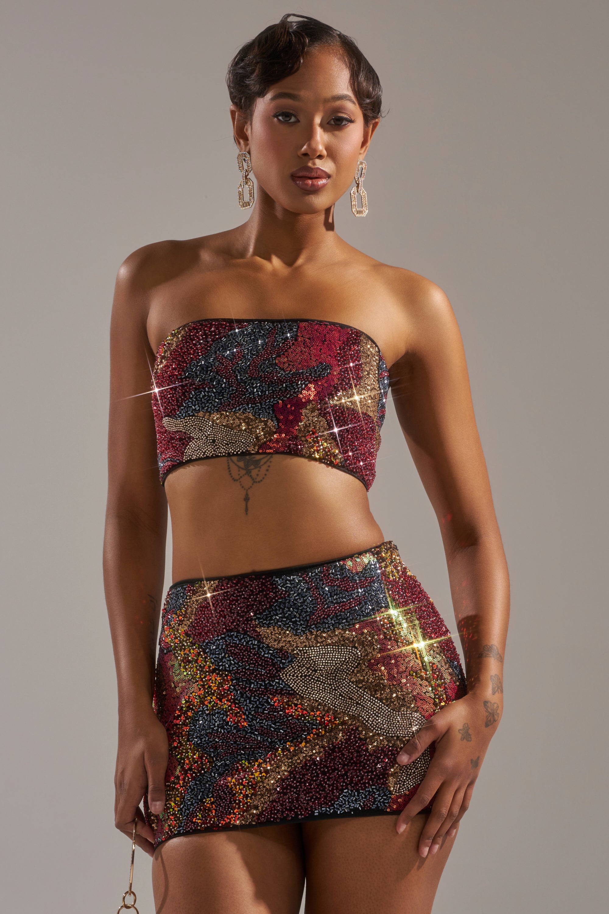 A woman with short styled hair and statement earrings poses confidently in the GOLDEN HOUR SEQUIN BRALETTE and matching mini skirt. She showcases arm and torso tattoos against a plain, neutral background. 1