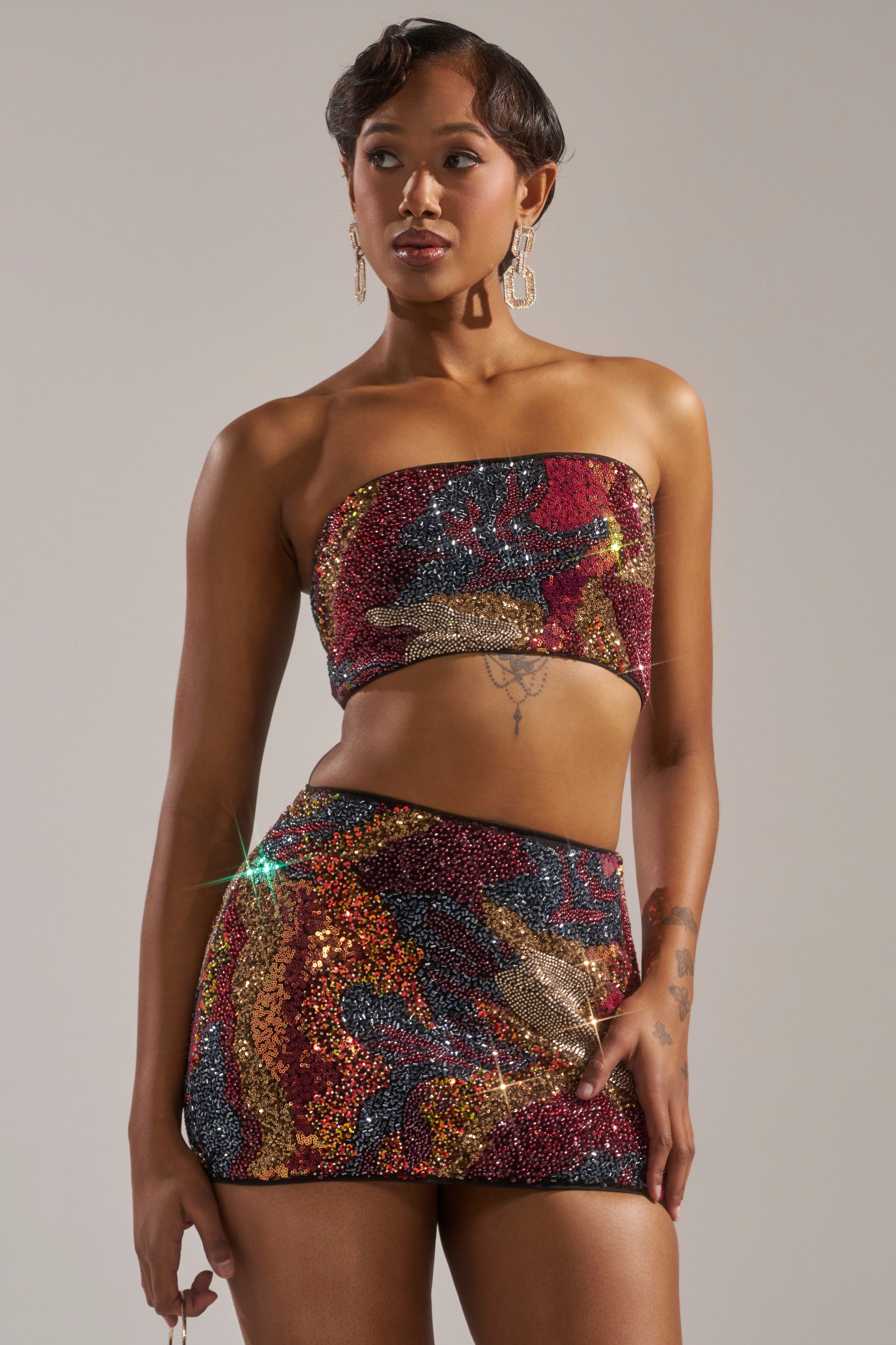 A woman models the GOLDEN HOUR SEQUIN BRALETTE and matching multicolored mini skirt, featuring a strapless, glittery design. She accessorizes with large earrings and showcases her short styled hair in glowing golden hour light. 3