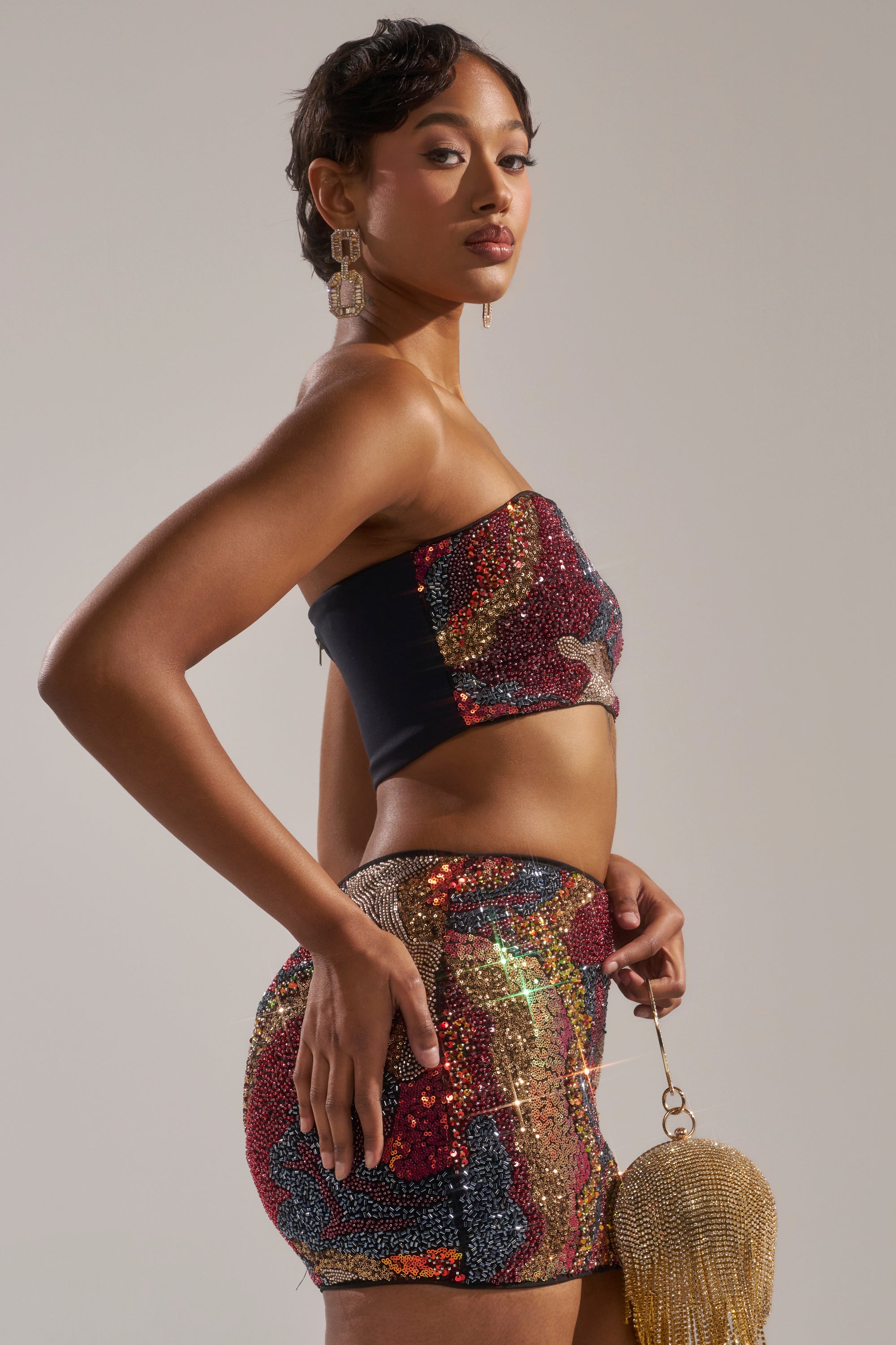 A woman with short hair and large earrings poses in the GOLDEN HOUR SEQUIN BRALETTE and matching mini skirt, holding a gold beaded clutch against a plain neutral background. 4