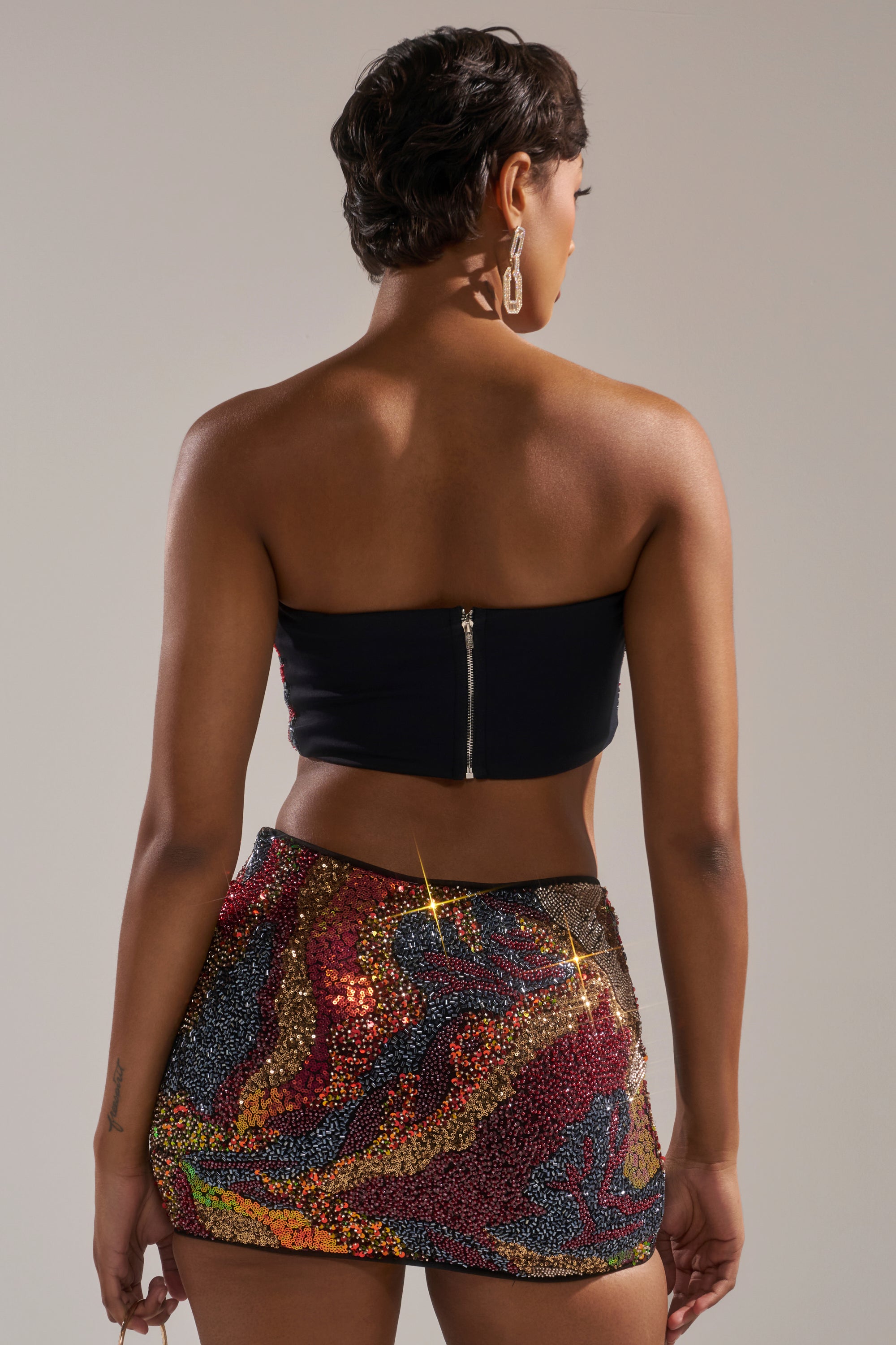 A woman with short hair and large earrings faces away, wearing the BLACK GOLDEN HOUR SEQUIN BRALETTE with a back zipper and a colorful sequined mini skirt, standing against a plain background. 5