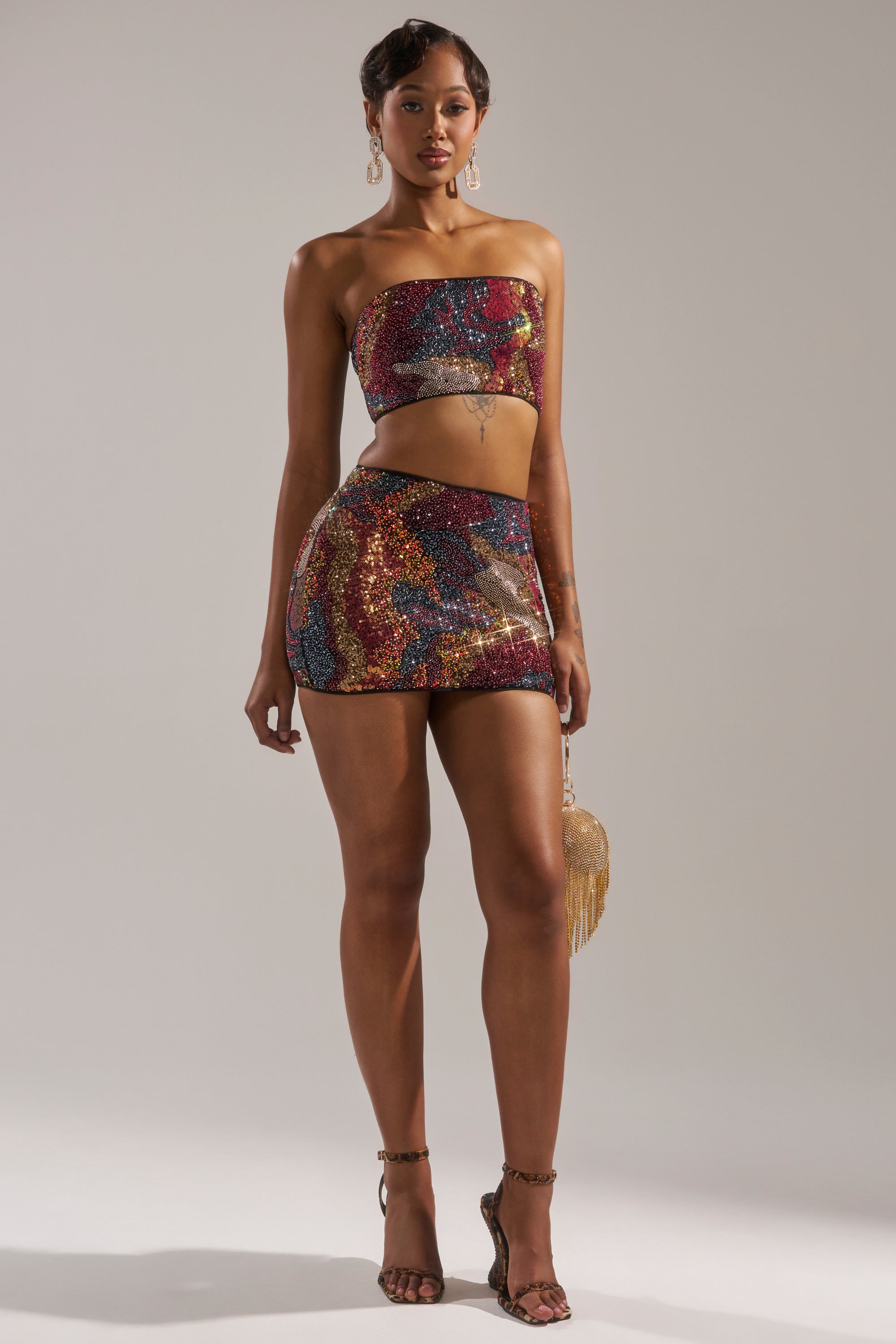 A woman stands confidently in the GOLDEN HOUR SEQUIN BRALETTE and matching mini skirt, holding a gold fringe purse. She completes her look with dangling earrings and strappy heels against a plain background. 8