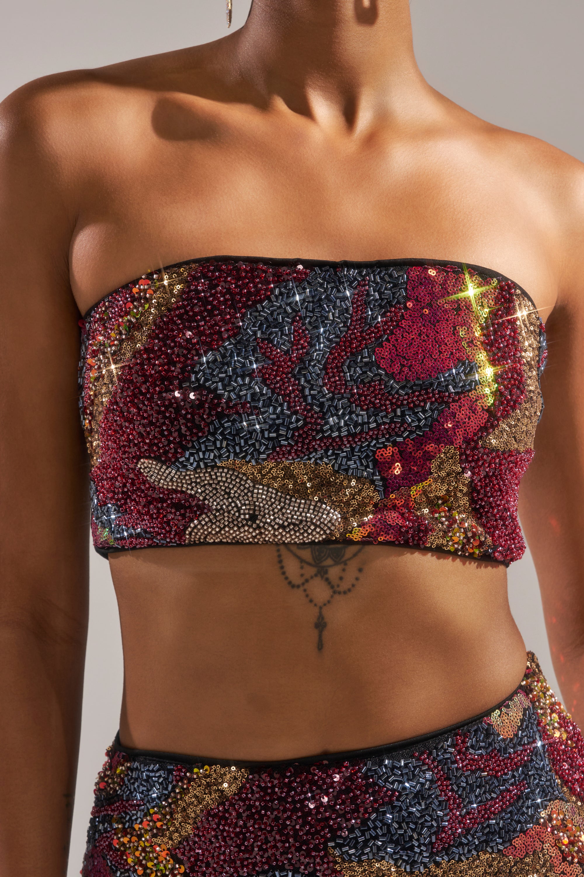 A person wears the GOLDEN HOUR SEQUIN BRALETTE with a vibrant red, gold, blue, and black abstract pattern. The photo captures their upper torso and highlights a delicate chest tattoo. 9