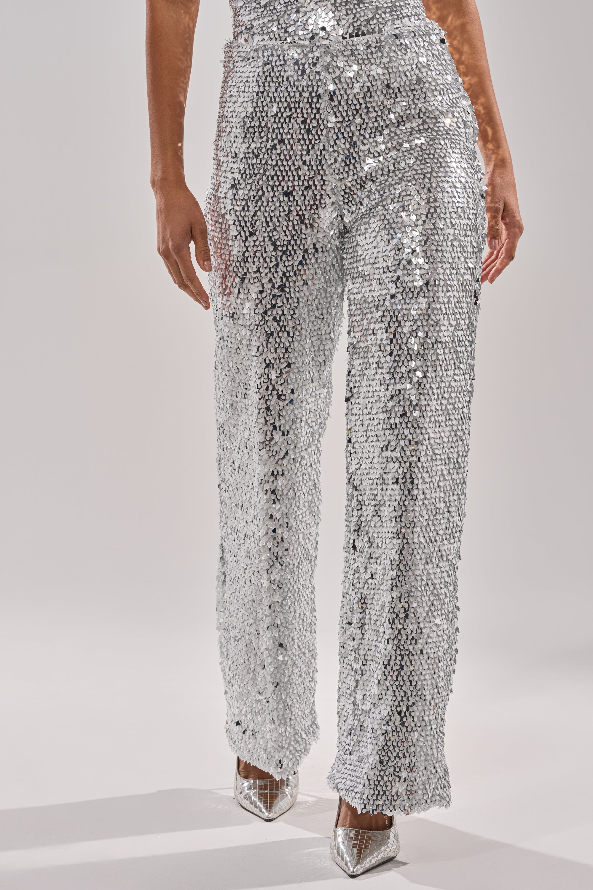 Wearing the NYE GIRLY TROUSER, a person showcases metallic sequin high-rise wide-leg pants and pointed transparent heels against a plain light background, with only their lower half visible. 0