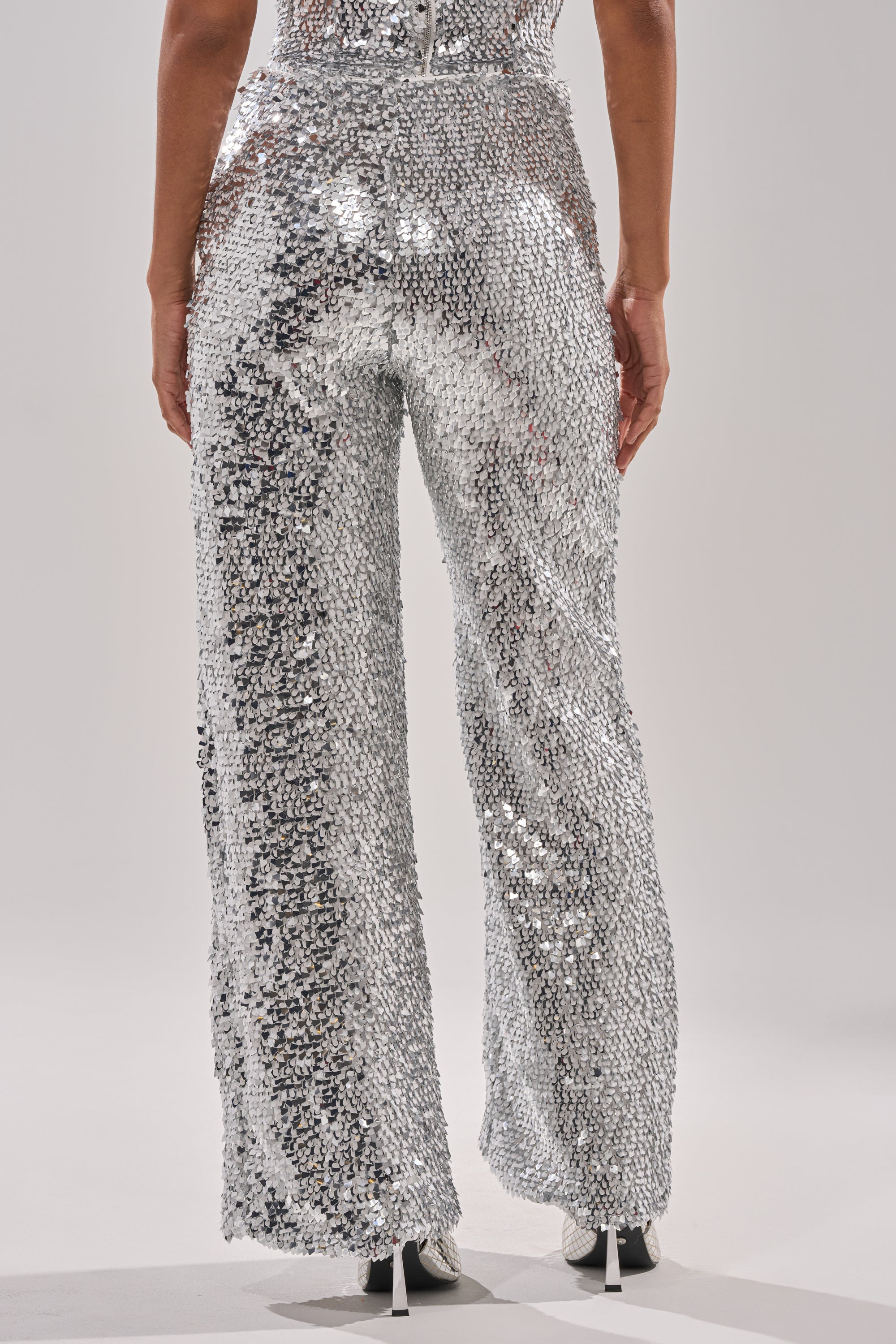 Facing away against a light background, a person wears the NYE GIRLY TROUSER—high-waisted, metallic sequin pants that sparkle brilliantly and reflect light with every move, paired with white high-heeled shoes. 4