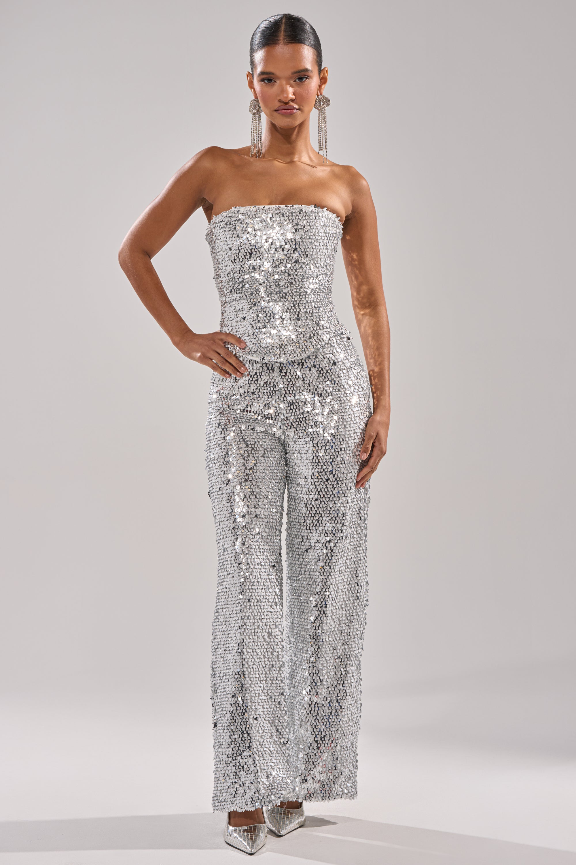 A woman stands confidently in NYE GIRLY TROUSER, a strapless silver sequin jumpsuit with wide-leg pants, styled with metallic pointed-toe heels and long statement earrings against a plain light background. 5