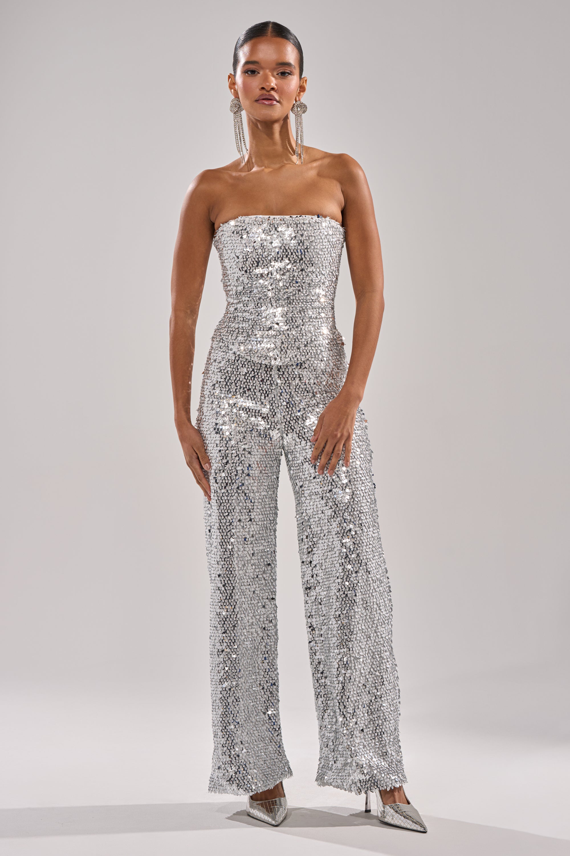 A woman stands confidently against a plain light gray background, wearing the NYE GIRLY TROUSER—a strapless silver metallic sequin jumpsuit with wide-leg mesh trousers, silver pointed-toe heels, and long dangling earrings. 6