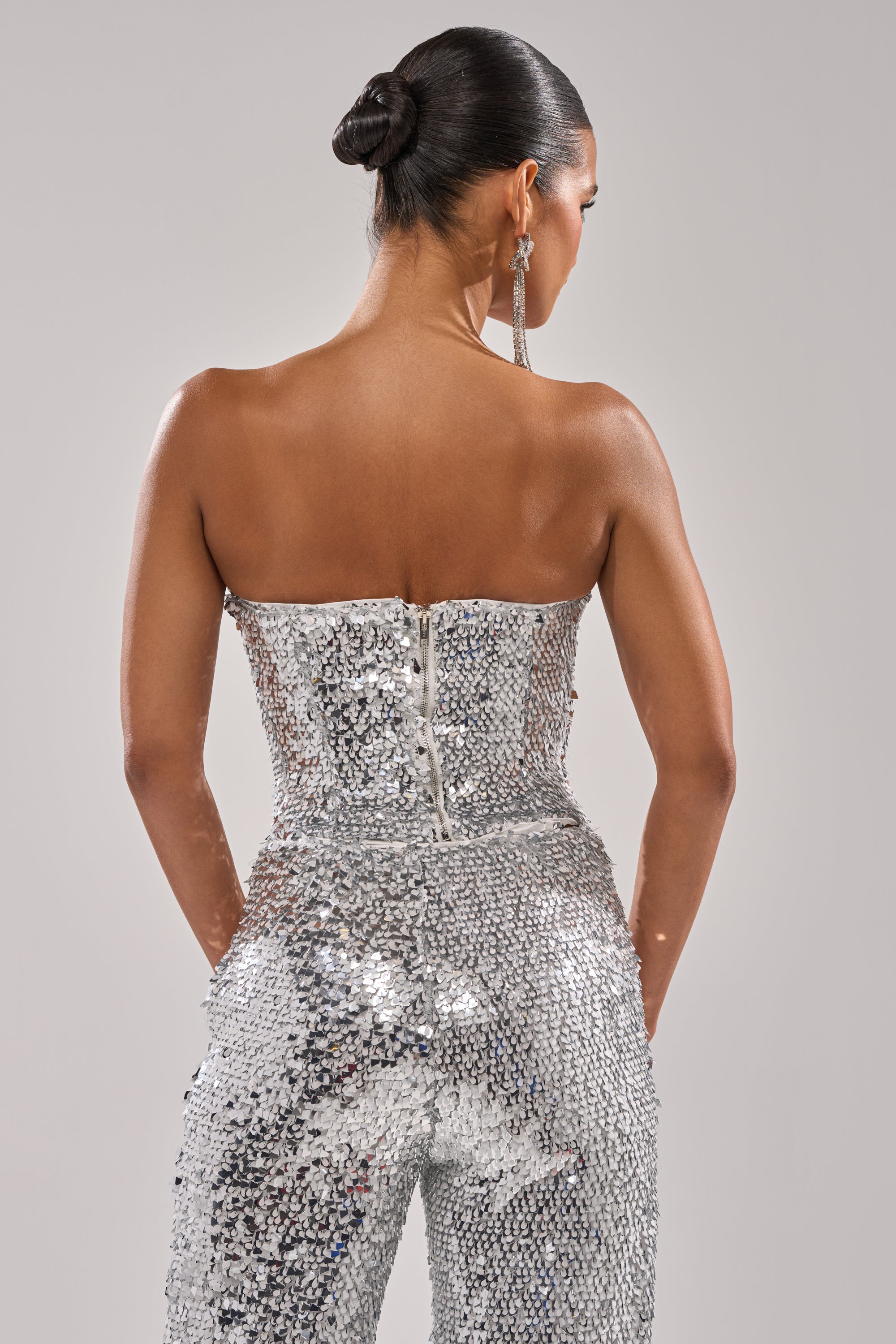 A woman with a sleek bun, seen from the back against a plain light gray background, wears the NYE GIRLY CORSET— a strapless mesh top paired with a sequined silver jumpsuit, her hands resting at her sides.