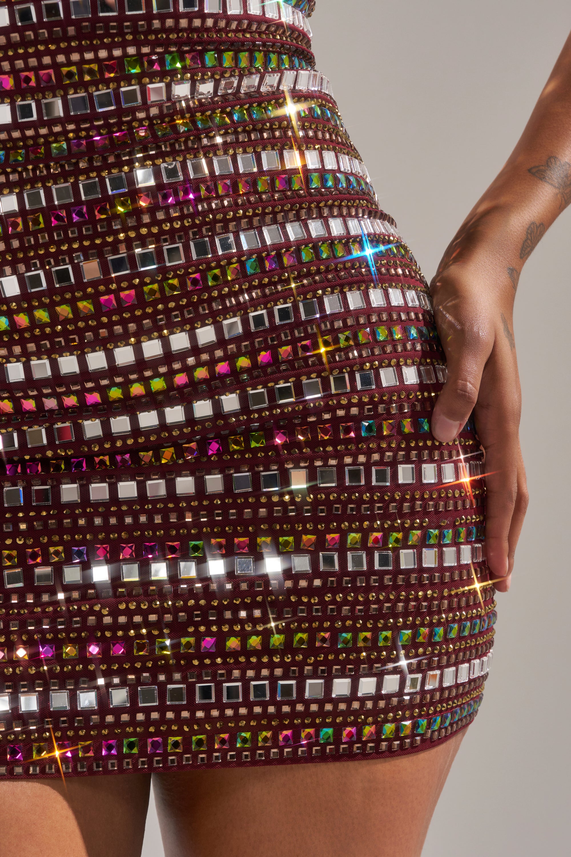 A close-up of a person wearing the LOOK AT HER GLOW RHINESTONE MINI DRESS, a fitted maroon mini dress with colorful rhinestone and sequin details. One hand is on their hip against a neutral background. 13