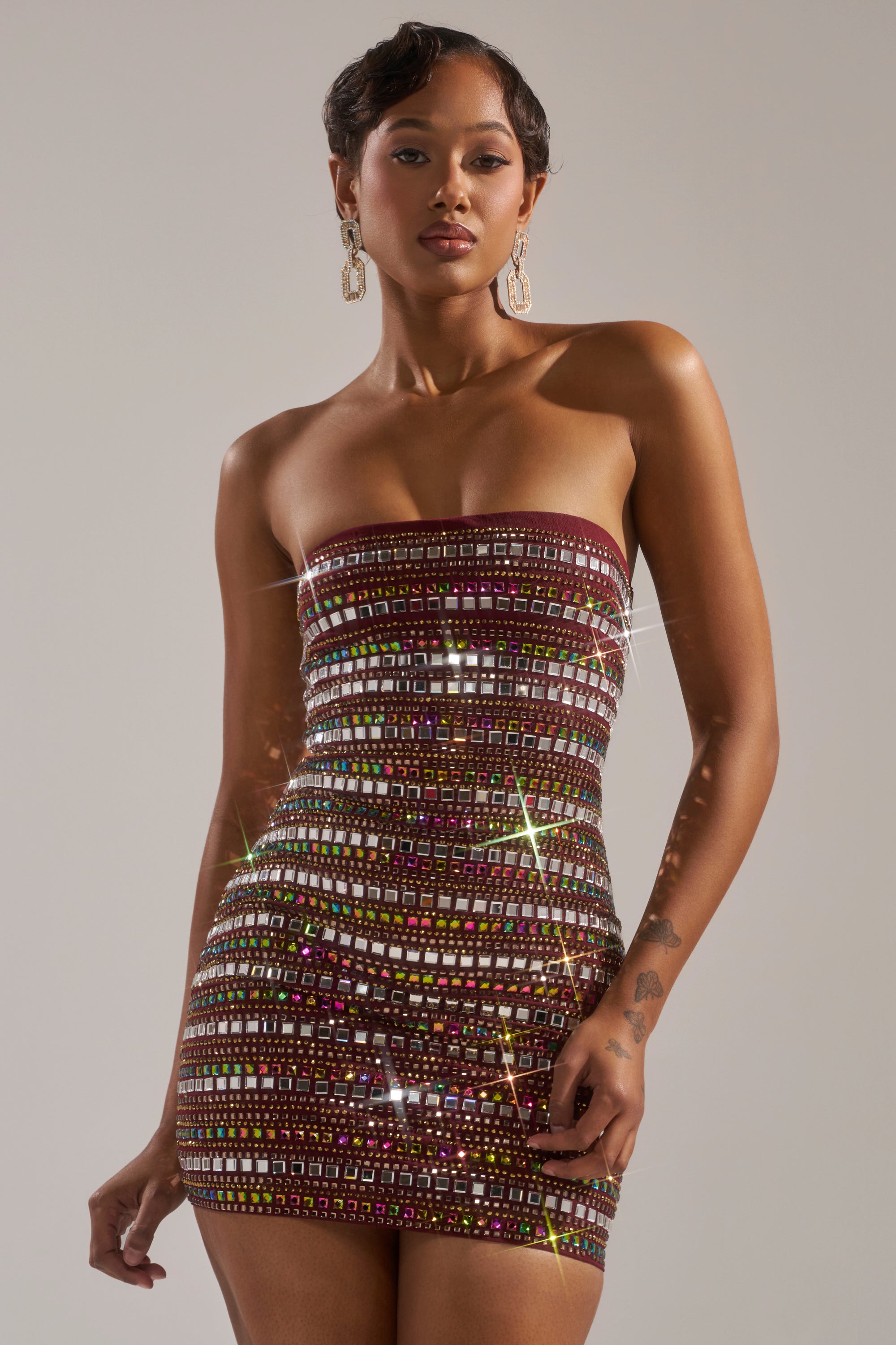 Wearing the LOOK AT HER GLOW RHINESTONE MINI DRESS, a woman poses against a gray background. The curve-hugging mini dress sparkles with multicolored sequins, while bold statement earrings and her short, wavy hair complete the look. 5