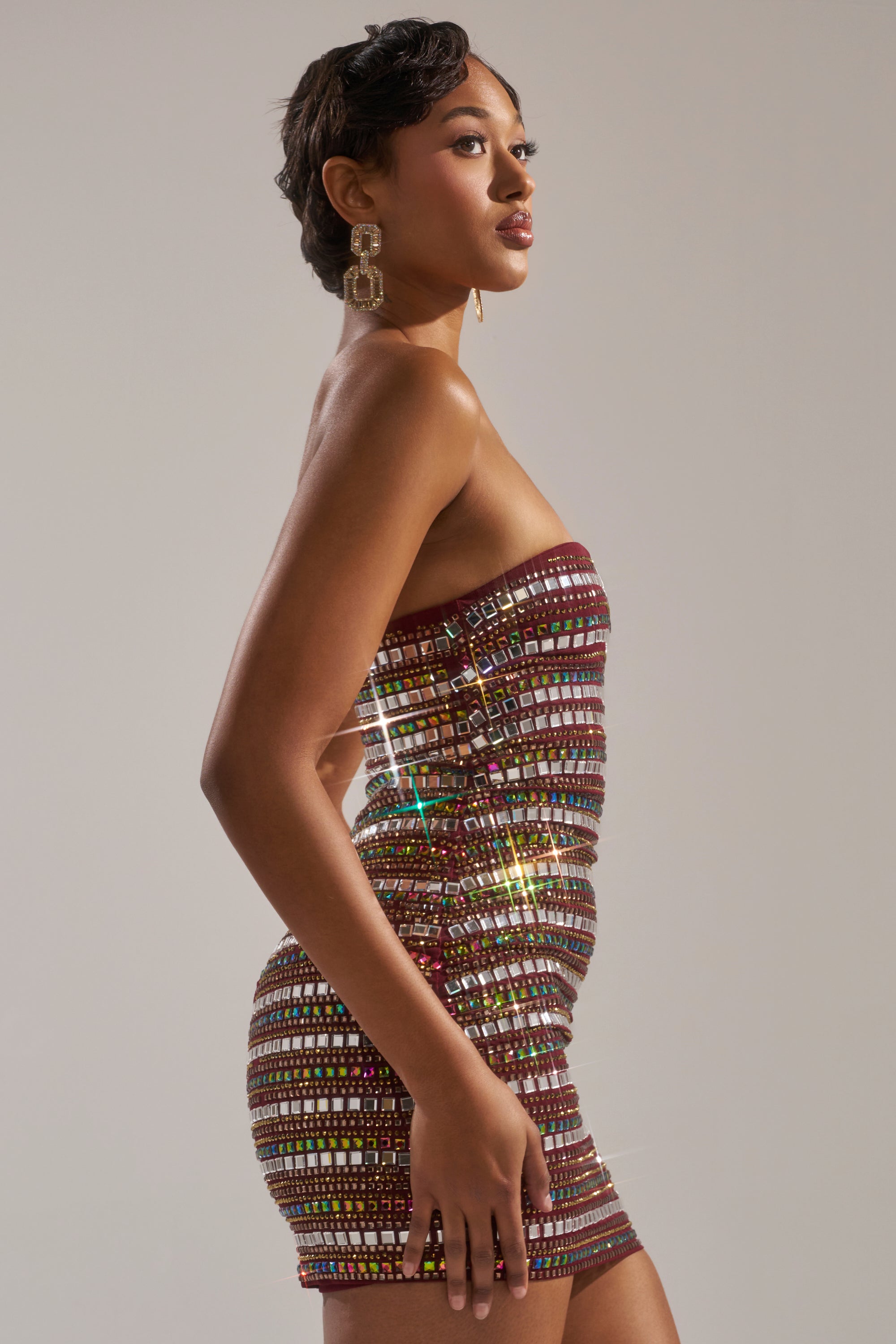 A woman poses in profile against a light background wearing the LOOK AT HER GLOW RHINESTONE MINI DRESS, a fitted, strapless mini dress adorned with multicolored sequins, paired with large rectangular gold earrings. 6