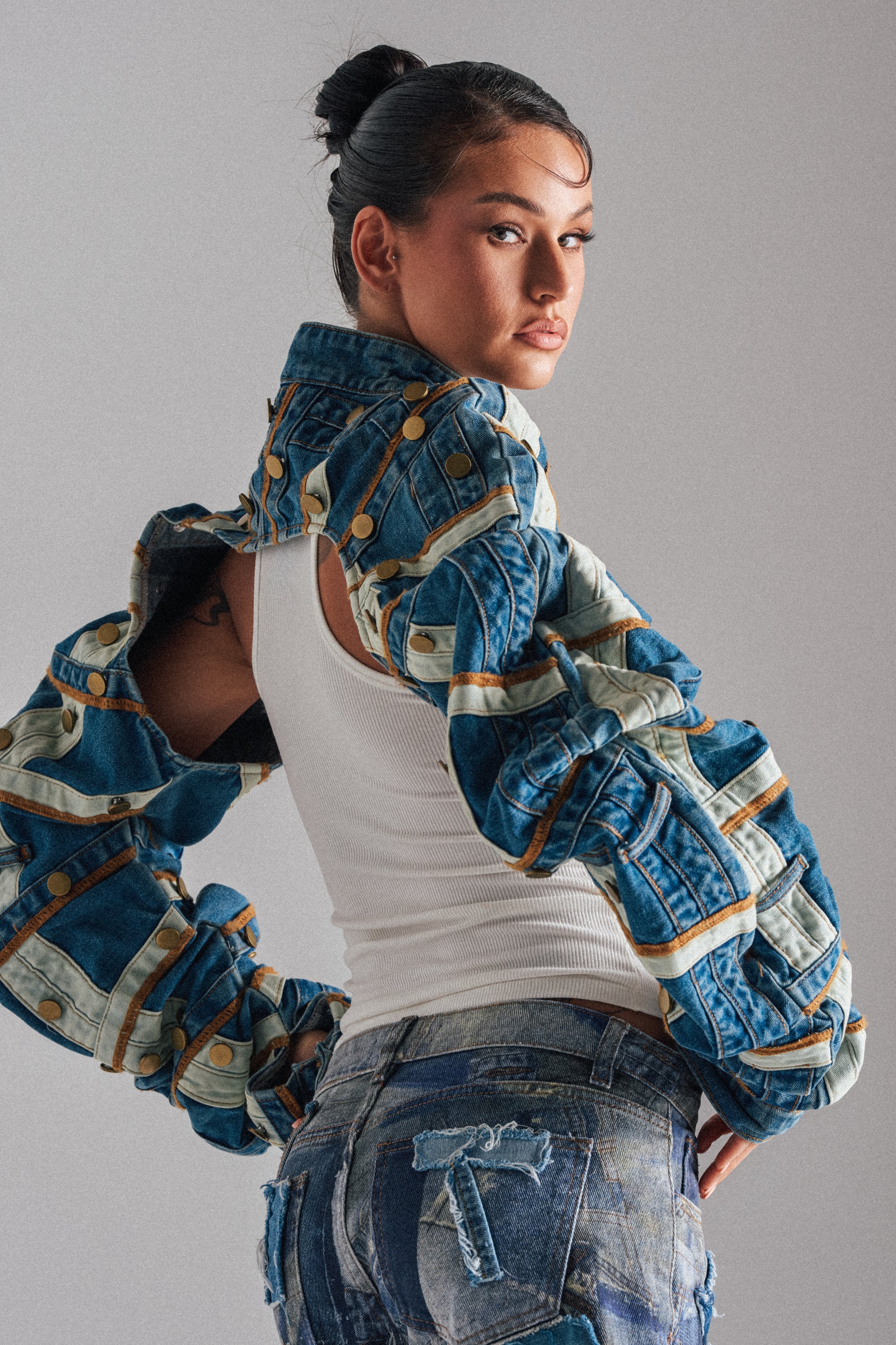 A woman with dark hair in a bun models the STACKED UP DENIM BOLERO, featuring exaggerated sleeves, over a white tank top and blue jeans with a denim patch. She poses against a plain background, looking over her shoulder. 1