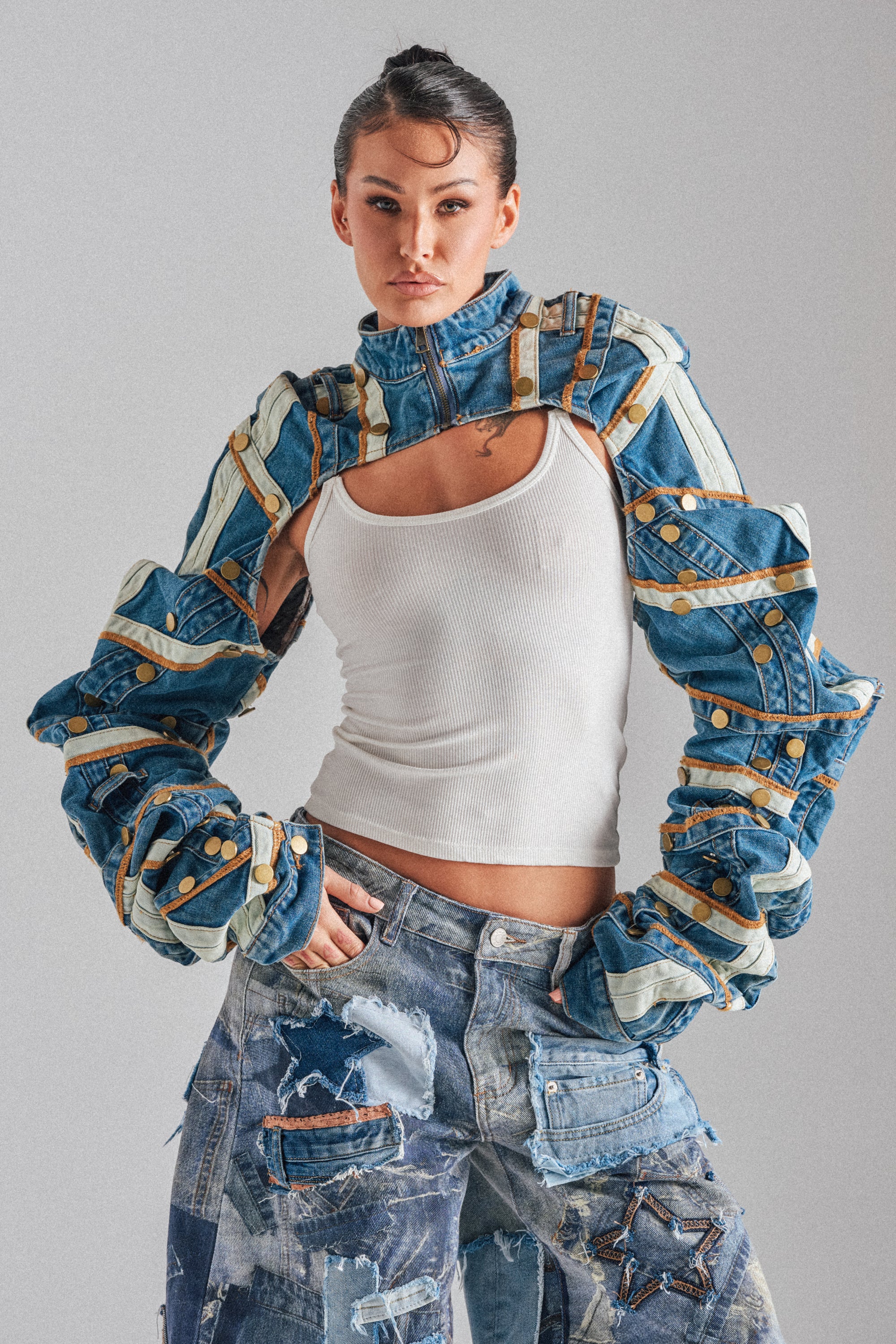 A woman models the STACKED UP DENIM BOLERO with cut-out sleeves over a white tank top and distressed patchwork jeans, standing against a plain gray background. 0