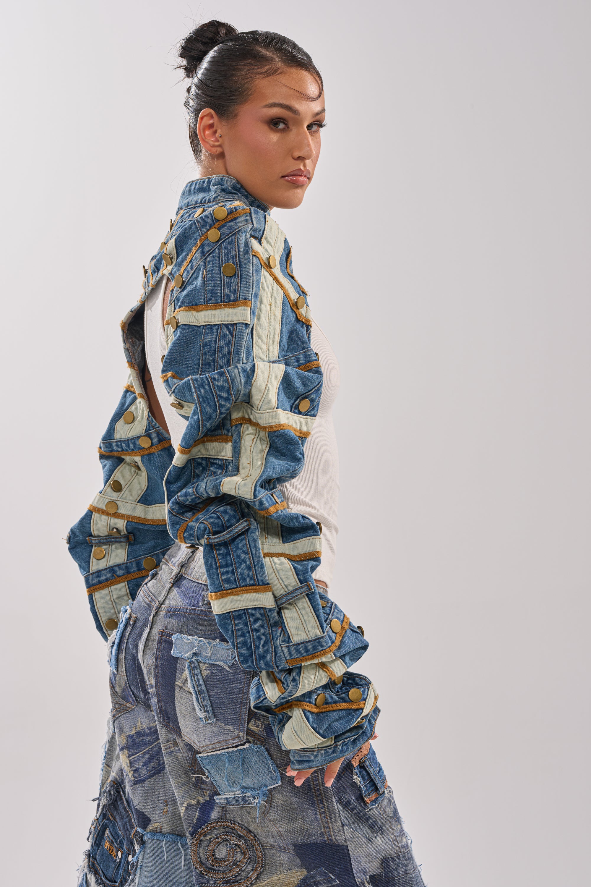 A woman with dark hair in a bun wears the STACKED UP DENIM BOLERO and a patchwork denim skirt, posing sideways against a plain light background. 5