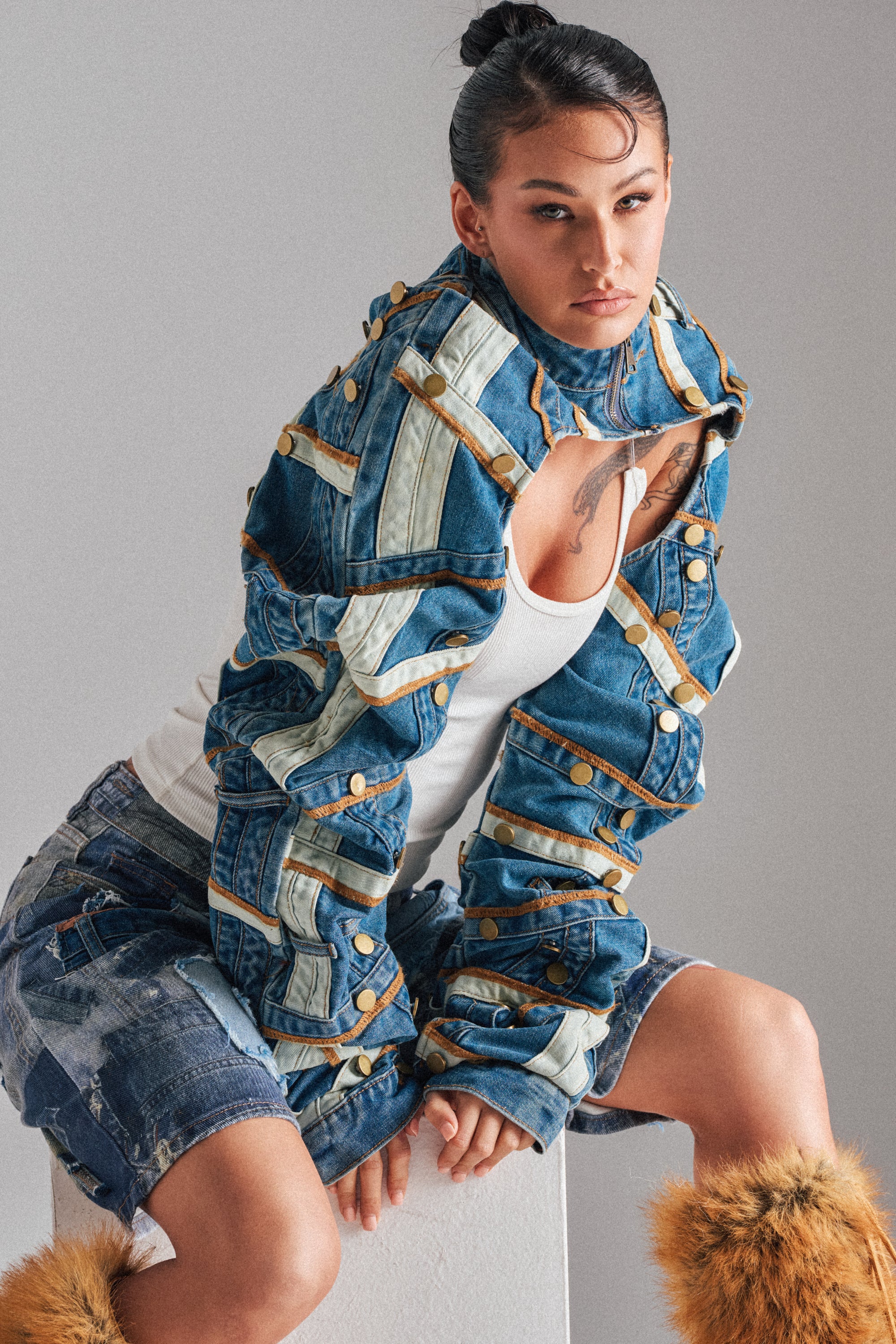 A woman poses confidently in the STACKED UP DENIM BOLERO—a unique patchwork denim piece with tan straps, buttons, and a cropped high-neck collar. She pairs it with a white tank top, matching shorts, and brown fluffy leg warmers. 2