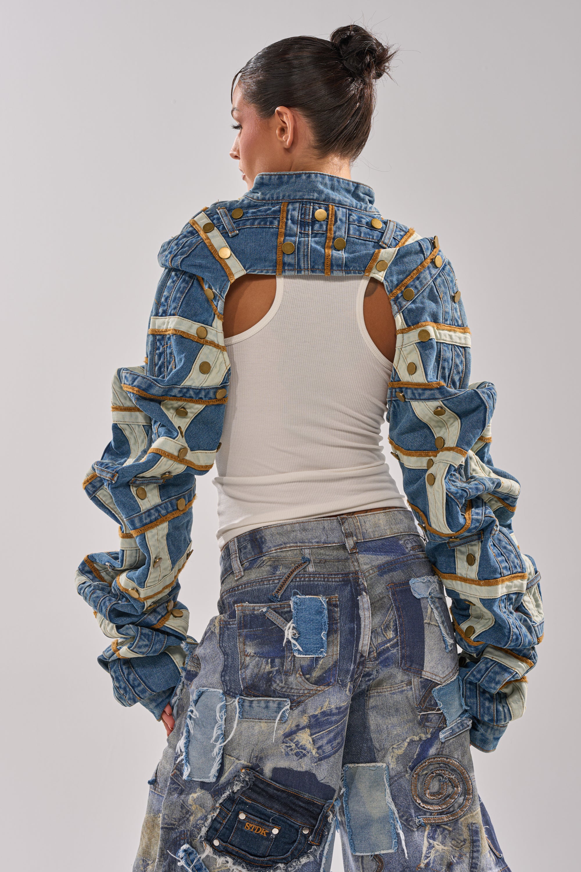 A person facing away wears a white tank top, the STACKED UP DENIM BOLERO with exaggerated ruched sleeves, and patchwork denim jeans in mixed shades and textures. Their hair is styled in a bun. 6