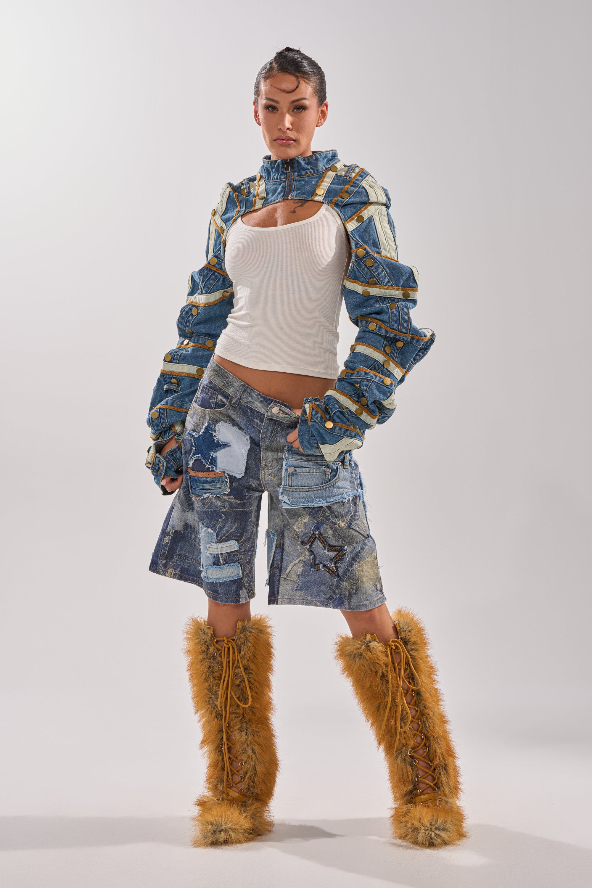 A woman poses in a white tank top, the STACKED UP DENIM BOLERO with wide sleeves, patchwork denim shorts, and tall furry orange boots against a plain light background. 7
