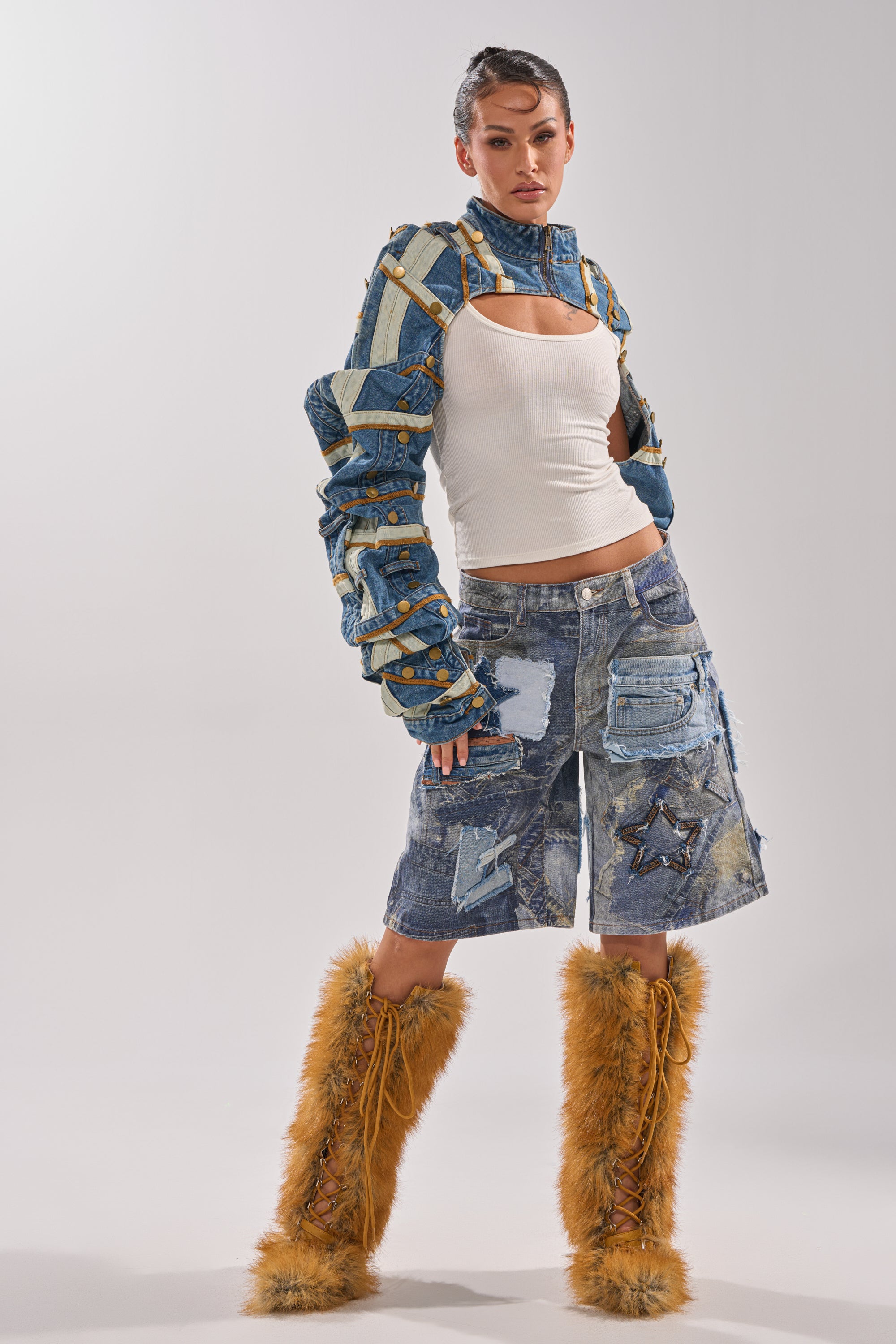 A woman poses against a white background wearing the STACKED UP DENIM BOLERO over a white tank, paired with a knee-length patchwork denim skirt and tall furry brown boots. 8