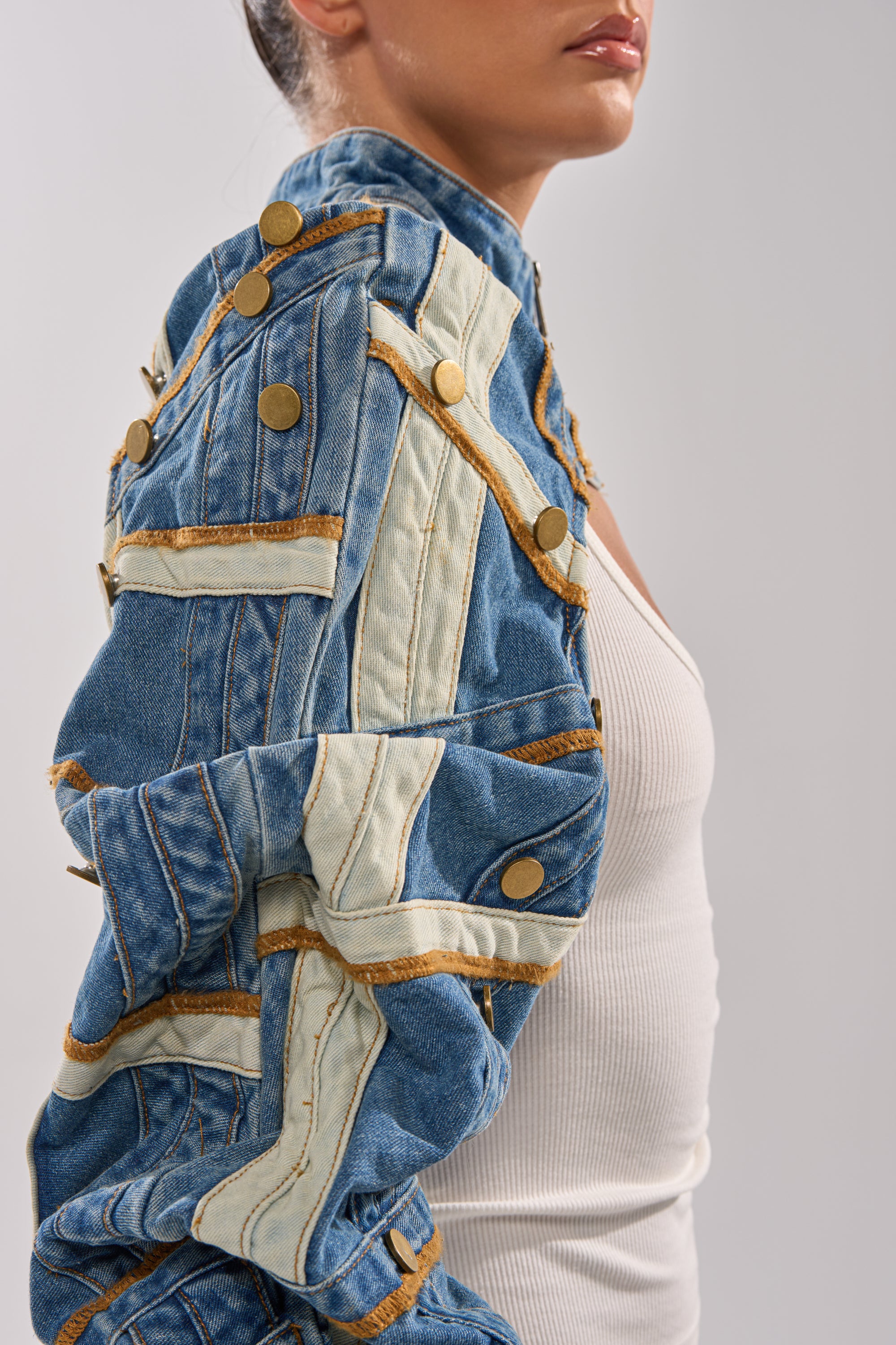 A woman wears the STACKED UP DENIM BOLERO—cropped with gold buttons and white-gold trim—over a white ribbed tank, photographed from the side against a plain, light background; her face is partially visible. 9