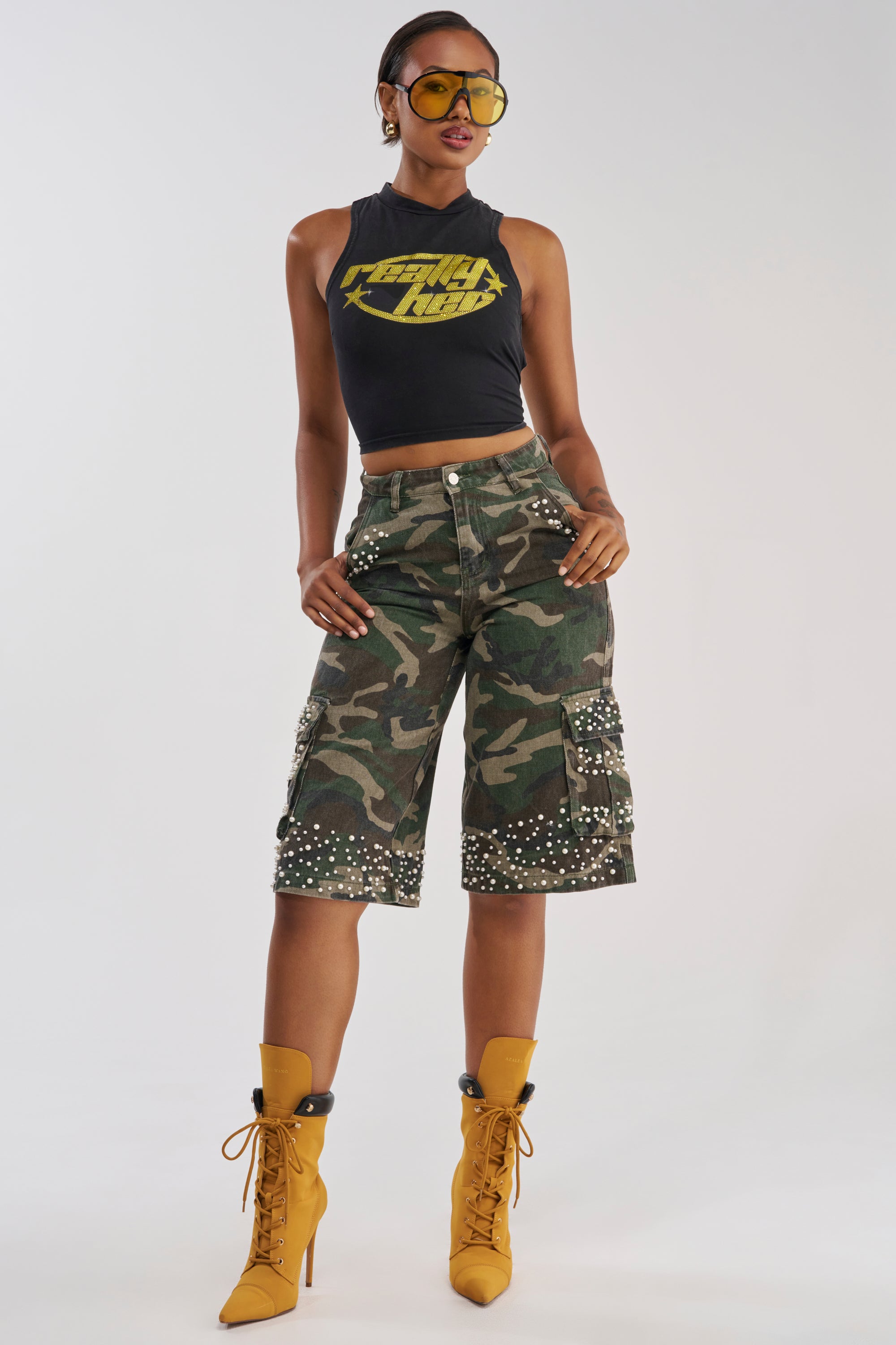 A woman poses against a plain white background wearing yellow sunglasses, a black sleeveless crop top, the LUXURIOUS CAMO BERMUDA CARGO SHORTS featuring faux pearl embellishments, and yellow lace-up boots.