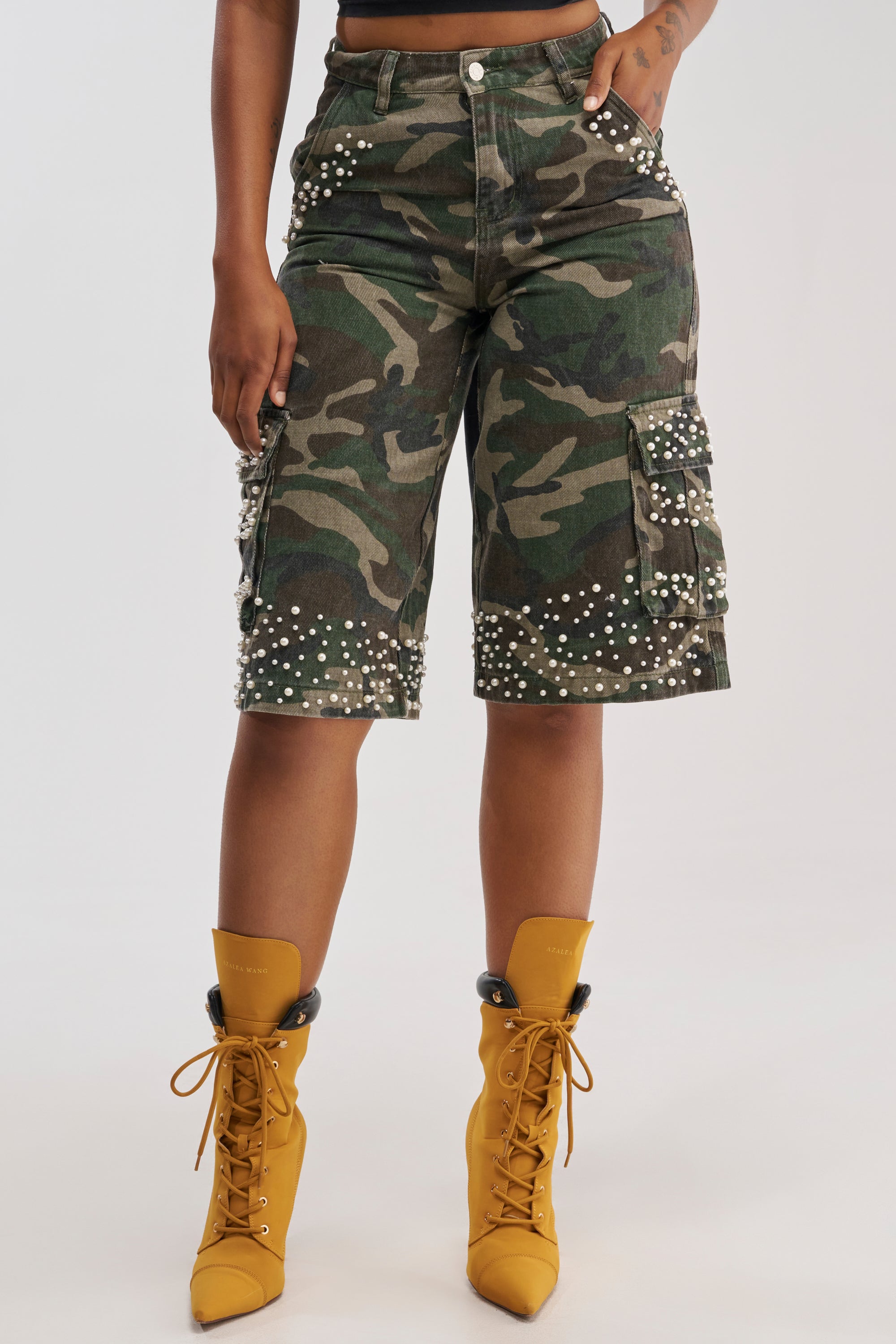 Wearing the LUXURIOUS CAMO BERMUDA CARGO SHORTS, a person stands against a plain background, their look completed with silver stud and faux pearl embellishments and yellow lace-up ankle boots.