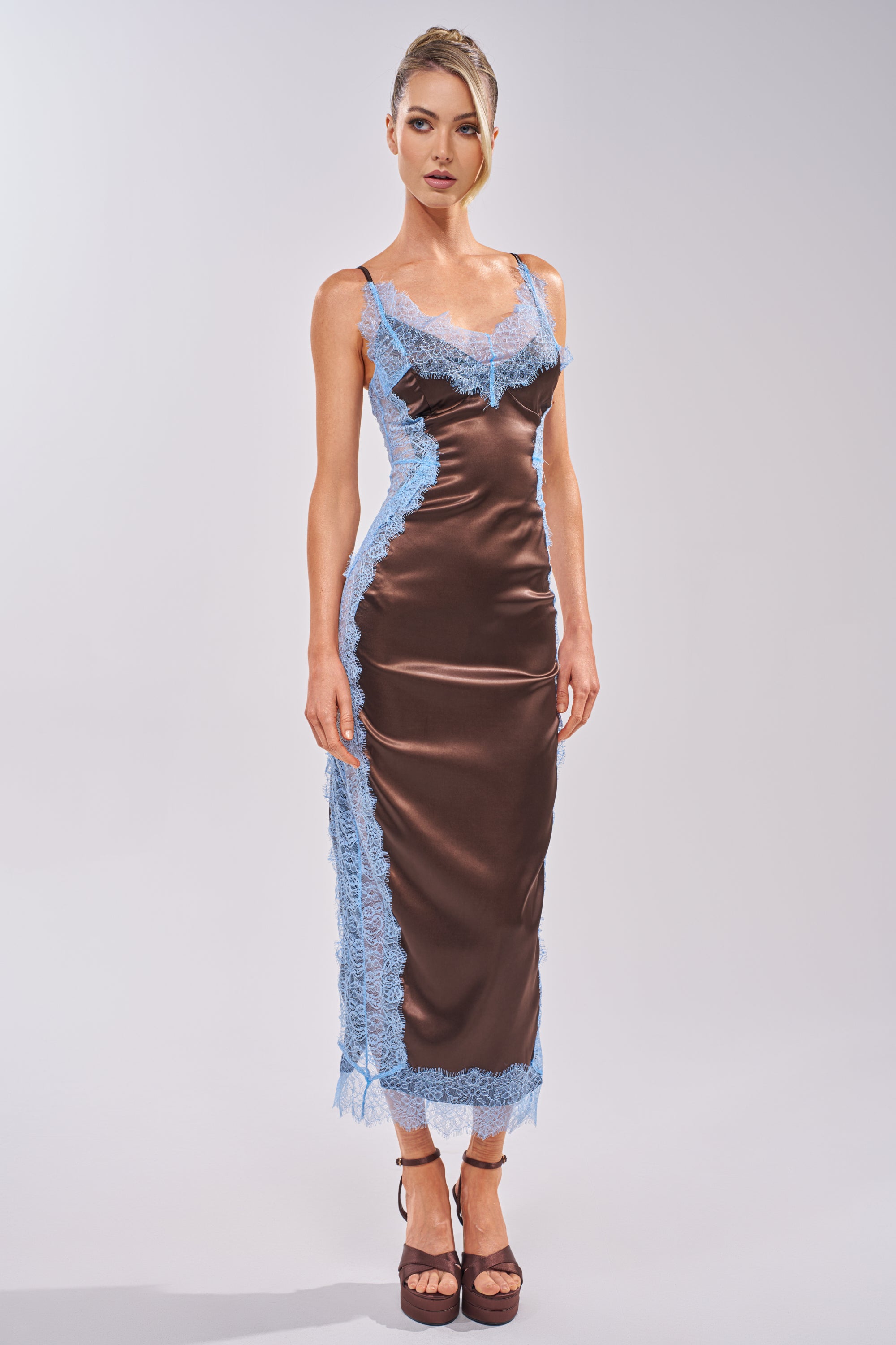 A woman with light hair in an updo and a neutral expression wears the ESSENTIAL LACE TRIM SATIN SLEEVELESS MAXI DRESS IN BROWN, featuring blue lace trim and thin straps, along with brown high-heeled sandals against a plain background. 1