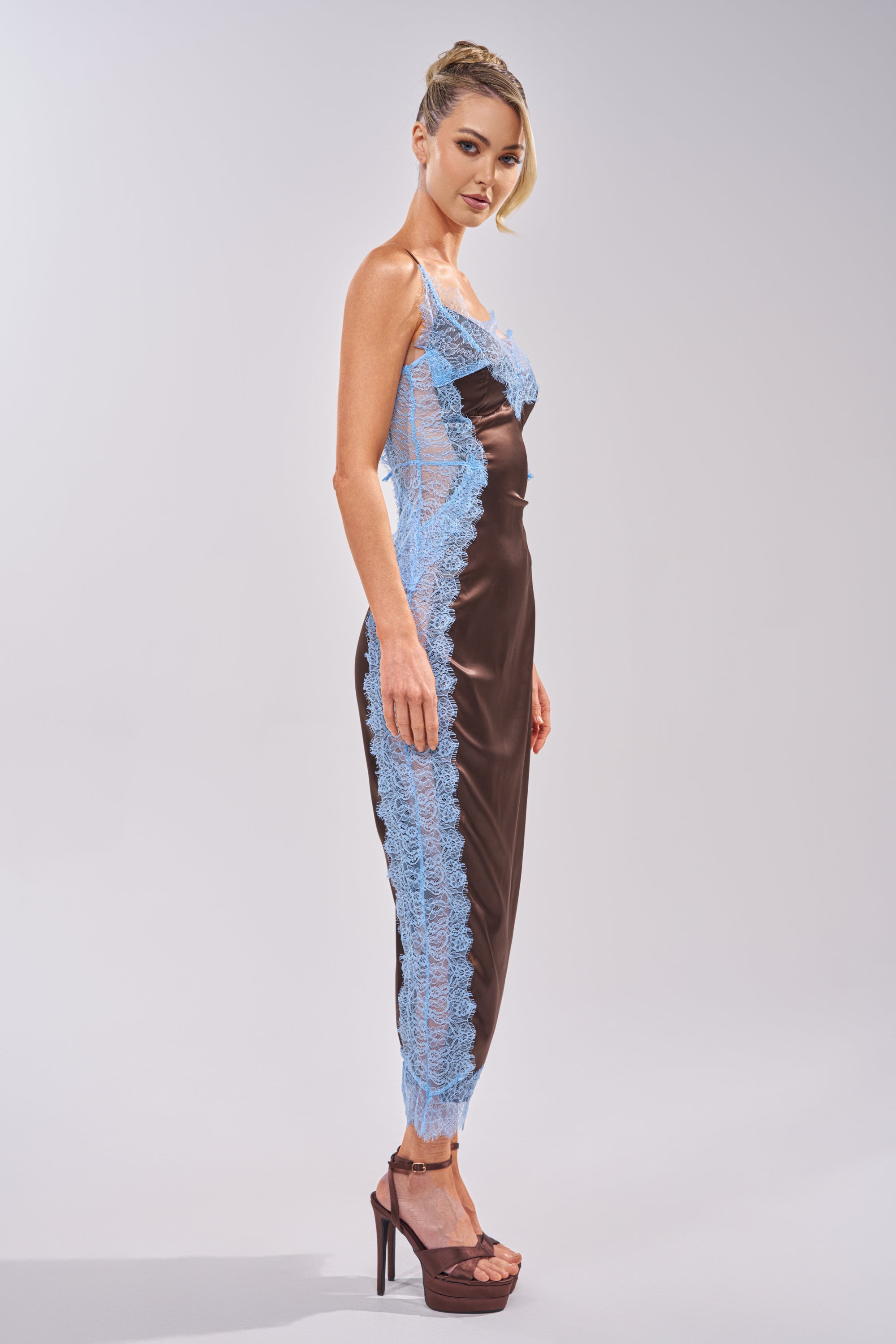 A woman in profile wears the ESSENTIAL LACE TRIM SATIN SLEEVELESS MAXI DRESS IN BROWN with blue lace trim, her blonde hair worn up. She pairs the look with high brown platform heels against a simple, light background. 2