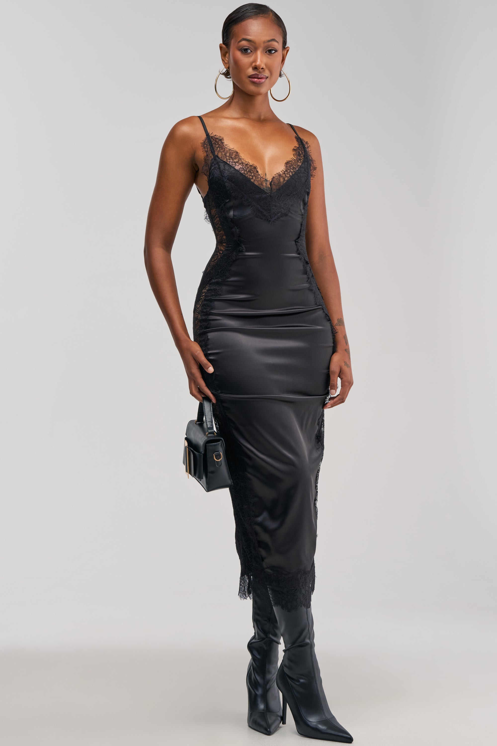 A woman wearing the ESSENTIAL LACE TRIM SATIN MIDI DRESS IN BLACK, styled with knee-high black boots, large hoop earrings, and a small black handbag, stands against a plain light background—an elegant choice for evening wear.