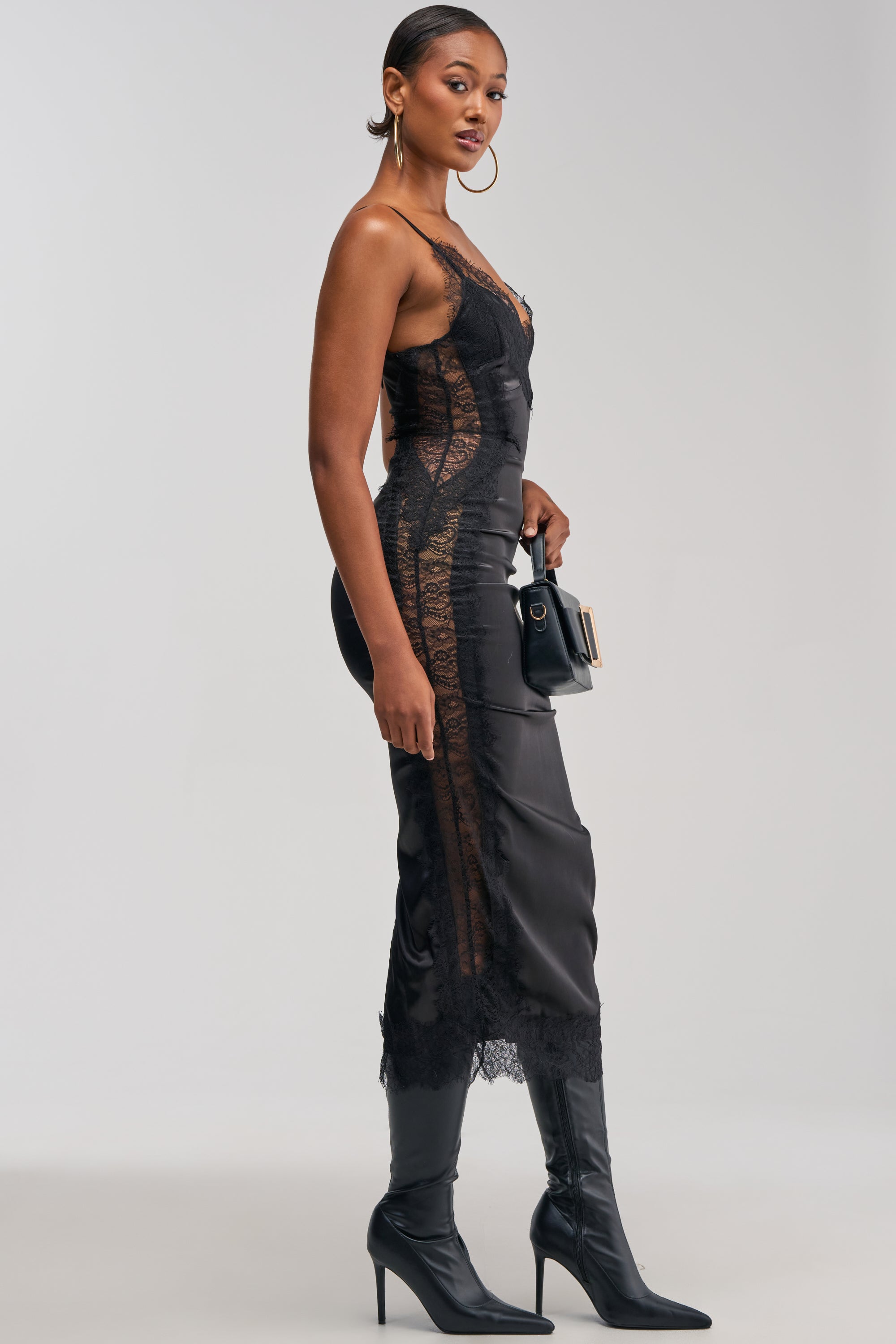 A woman wearing the ESSENTIAL LACE TRIM SATIN MIDI DRESS IN BLACK, styled with black heeled boots and gold hoop earrings, stands sideways holding a black purse against a plain background.