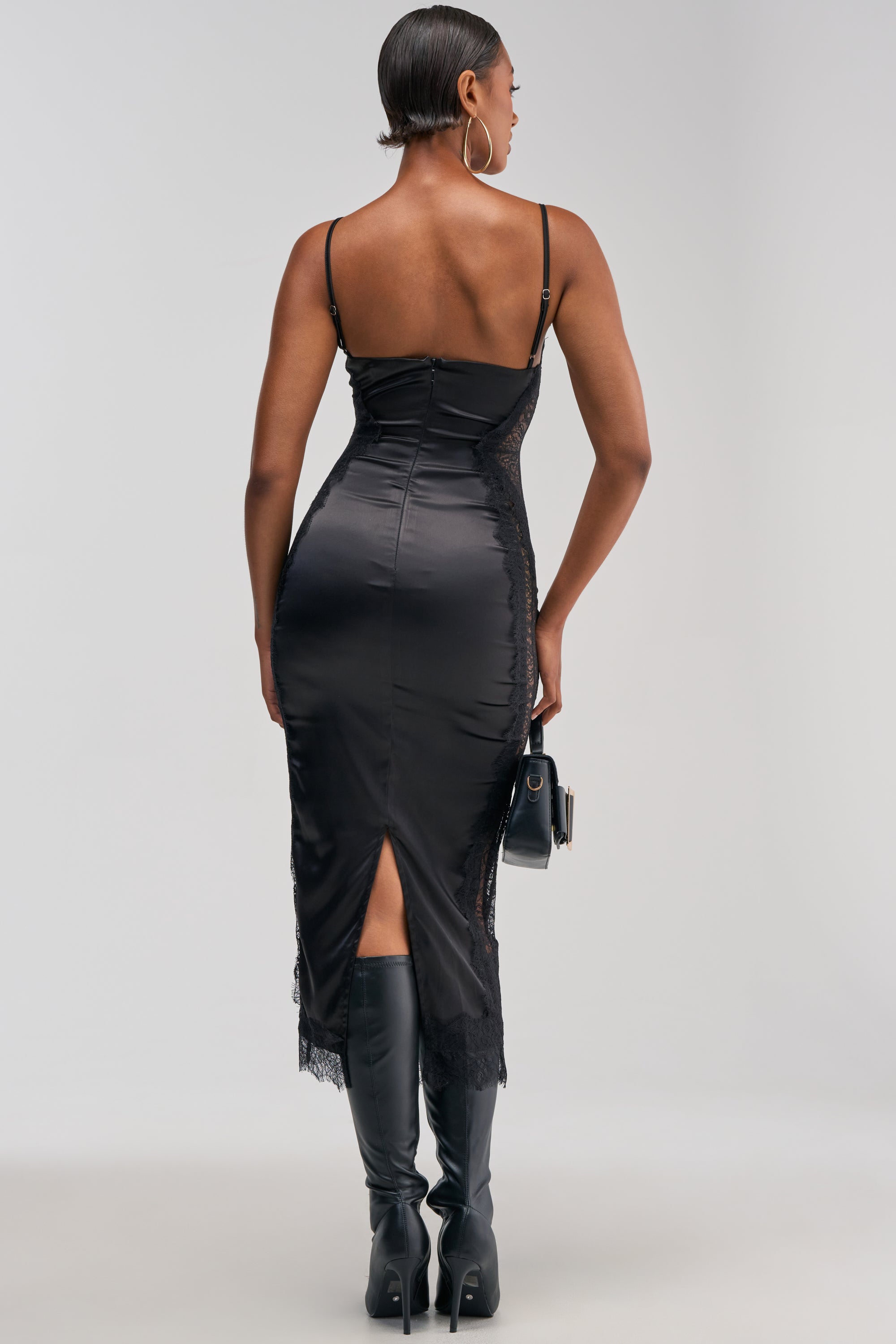 A woman stands facing away, wearing the ESSENTIAL LACE TRIM SATIN MIDI DRESS IN BLACK—an elegantly fitted black satin midi dress featuring lace trim details and a back slit—styled with knee-high boots. Her hair is slicked back, and she holds a small handbag against a plain, light background.