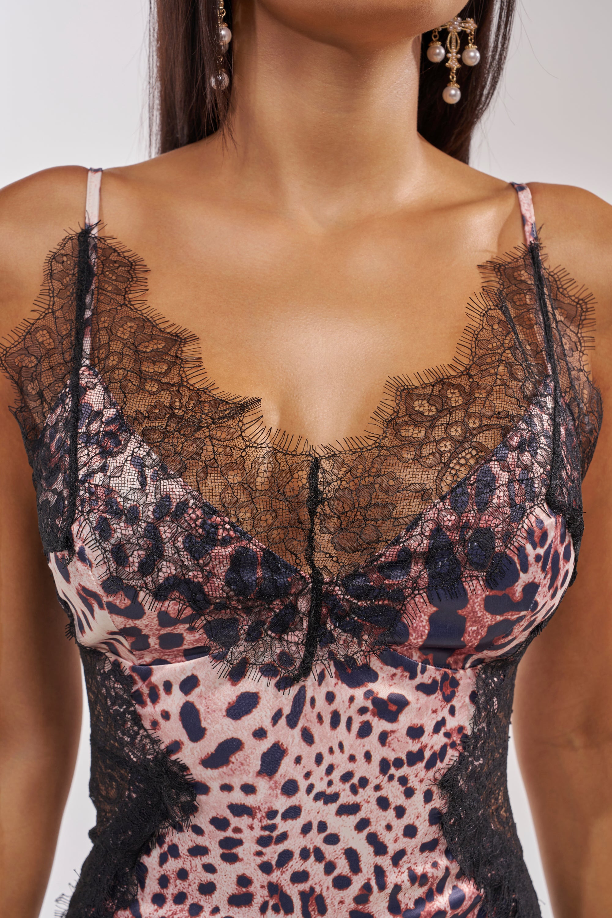 A close-up of a person wearing the SHOW US WHAT YOU GOT SATIN MIDI DRESS IN CHEETAH, a sleeveless cheetah-print dress with black lace-trimmed neckline. The person has long dark hair and is wearing dangling earrings. 6