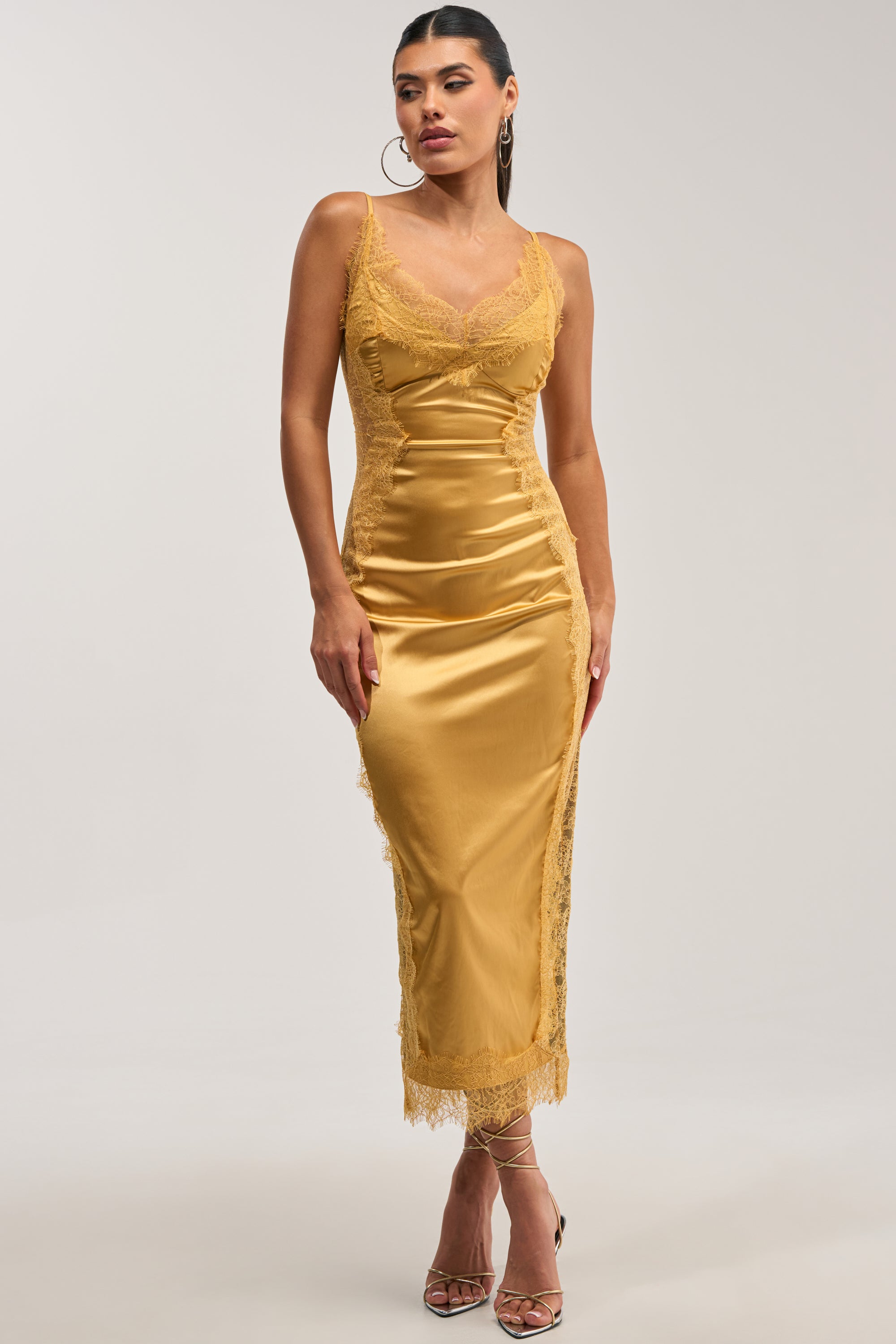 A model poses against a white background wearing the ESSENTIAL LACE TRIM SATIN SLEEVELESS MIDI DRESS IN GOLD, which features spaghetti straps and delicate lace trim along the neckline and side panels. She completes the look with thin-strapped gold heels and hoop earrings.